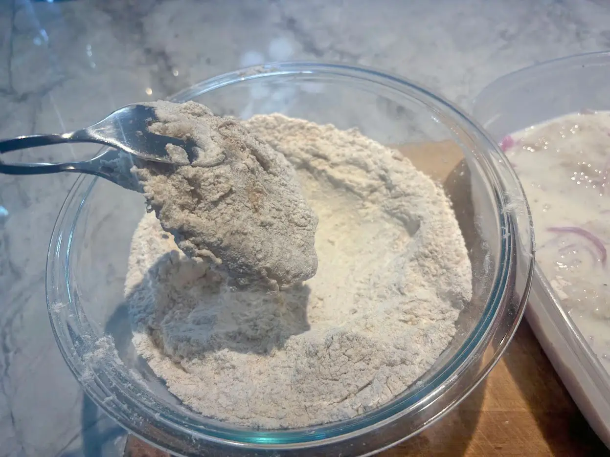 Tongs dipping raw chicken into the flour mixture.