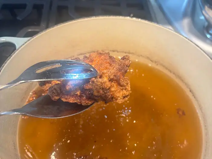 Tongs holding fried chicken over a Dutch oven filled with canola oil.