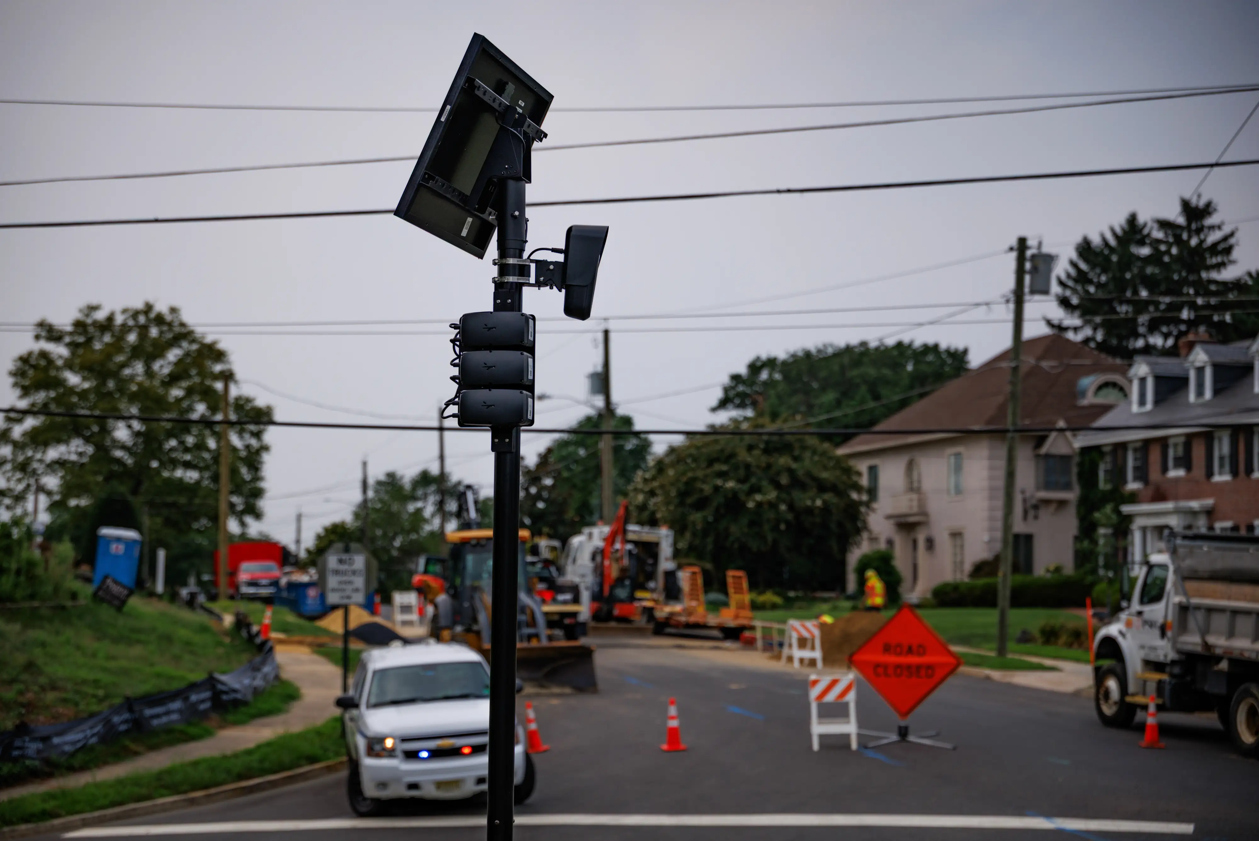 A Flock camera is mounted onto a black pole in a New Jersey town. There's a police cruiser in the background.