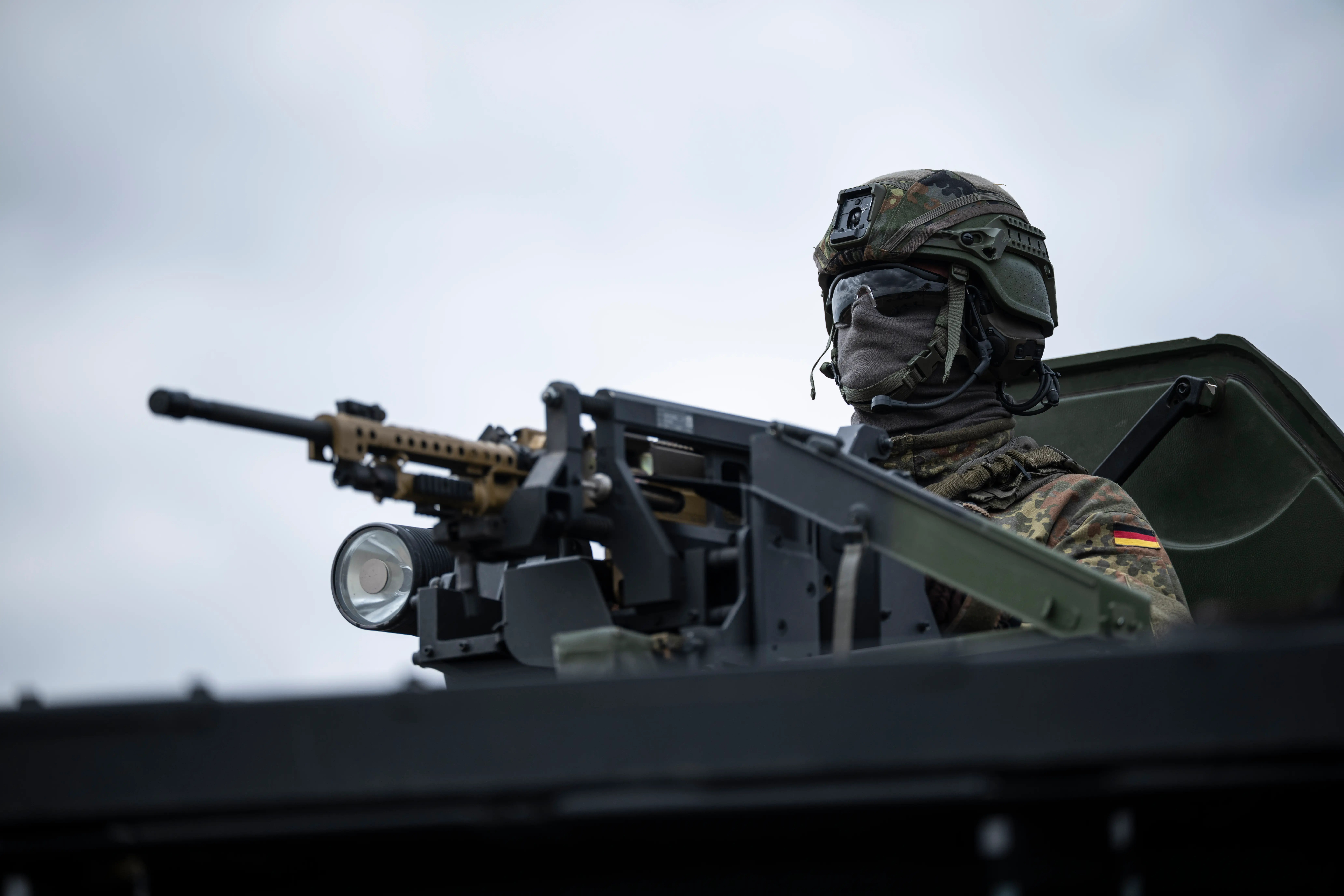 Camouflaged soldier with a German flag patch operates a mounted machine gun on an armored vehicle.