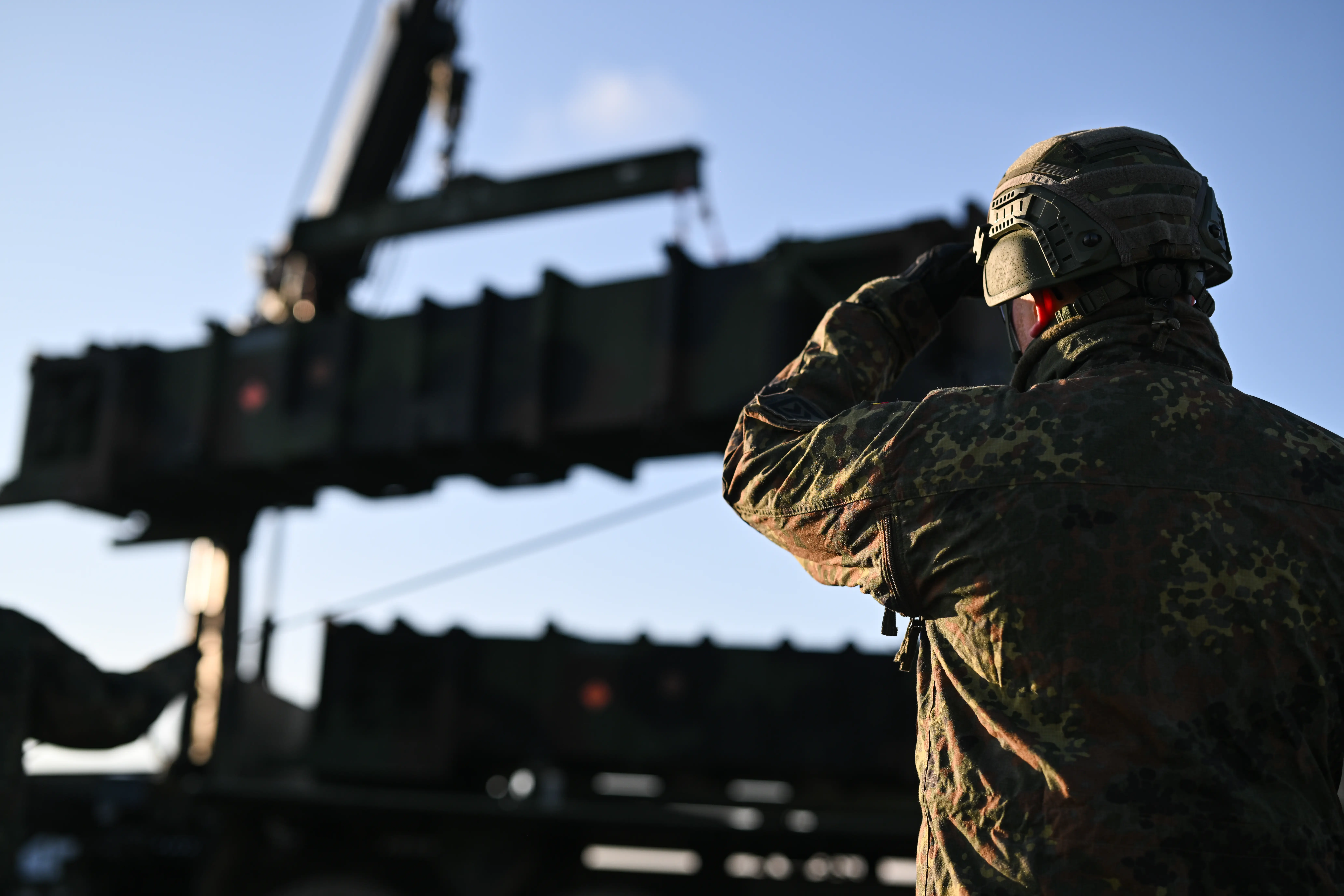 Camouflaged soldier stands beside heavy military equipment and a crane under a clear sky.