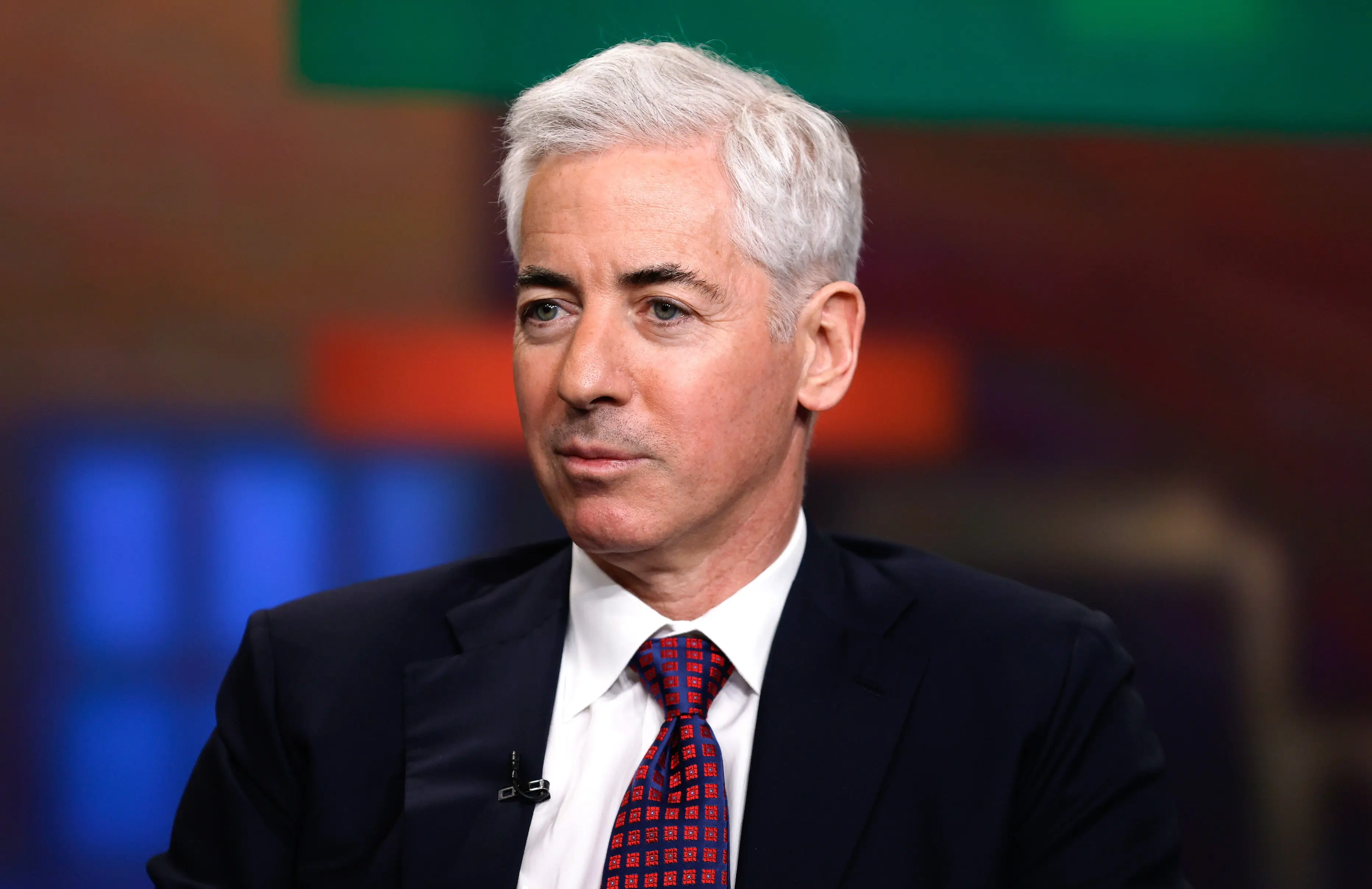 Bill Ackman sits in a studio wearing a dark suit, white shirt, red patterned tie, and lapel microphone.