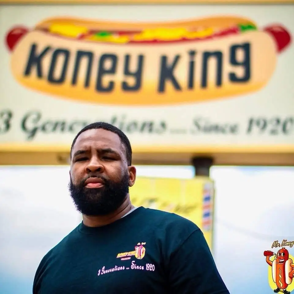 Jimmy Hendricks in a dark Koney King T-shirt stands in front of a large Koney King hot dog sign.