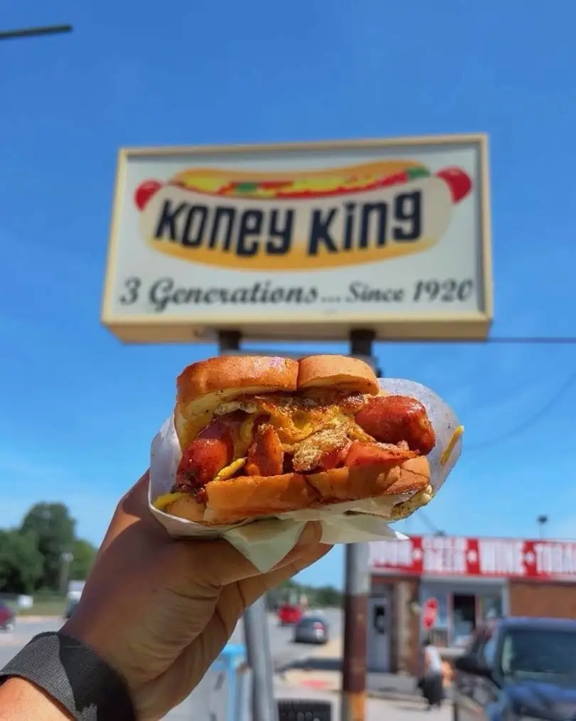 A hand holds a chili-style hot dog in front of a Koney King roadside sign under a blue sky.