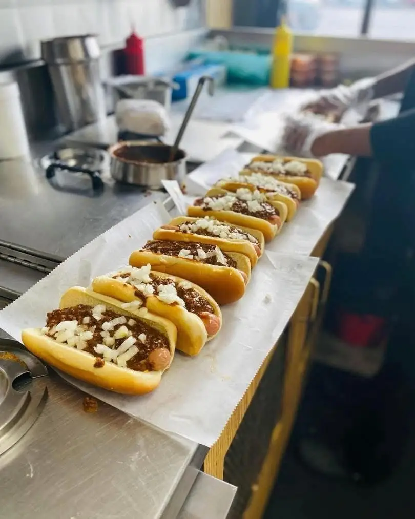Chili hot dogs topped with chopped onions line a parchment-covered counter in a food preparation area.