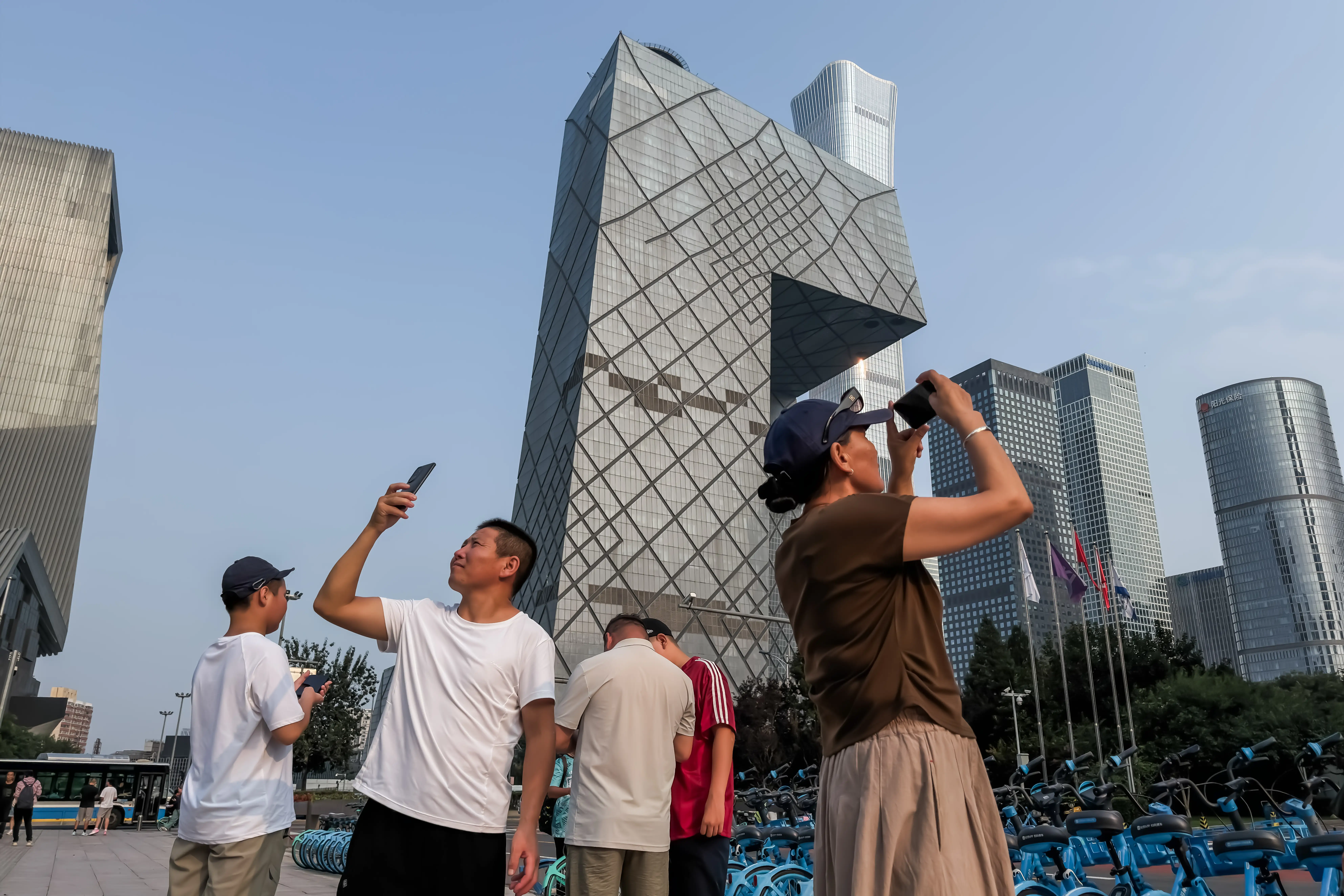 People take photographs with smartphones near the China Central Television (CCTV) Headquarters as CITIC Tower, also known as China Zun, rises in the background on August 22, 2026, in Beijing, China.