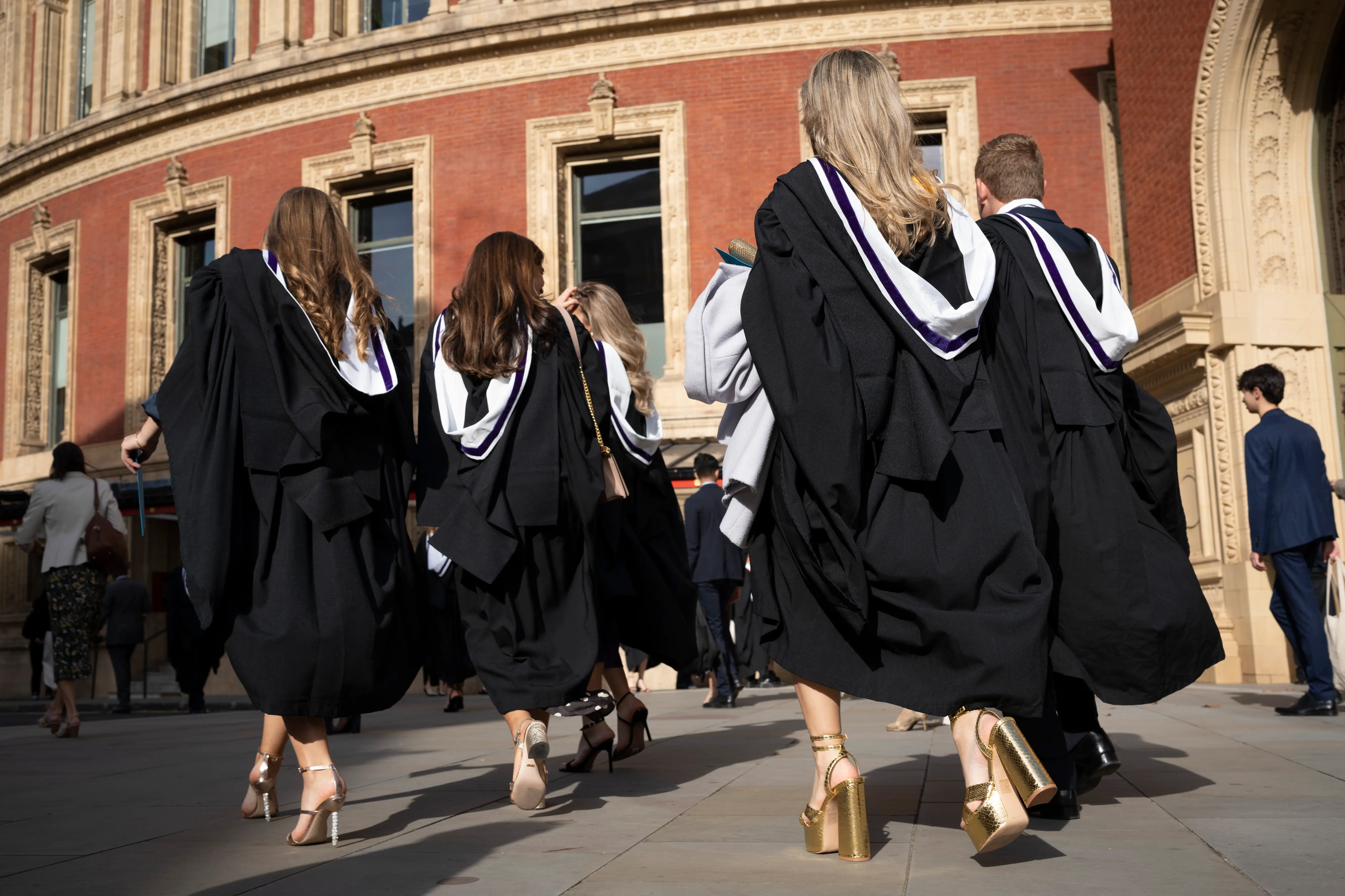 Graduates in black academic gowns walk toward an ornate red brick building entrance in bright sunlight.