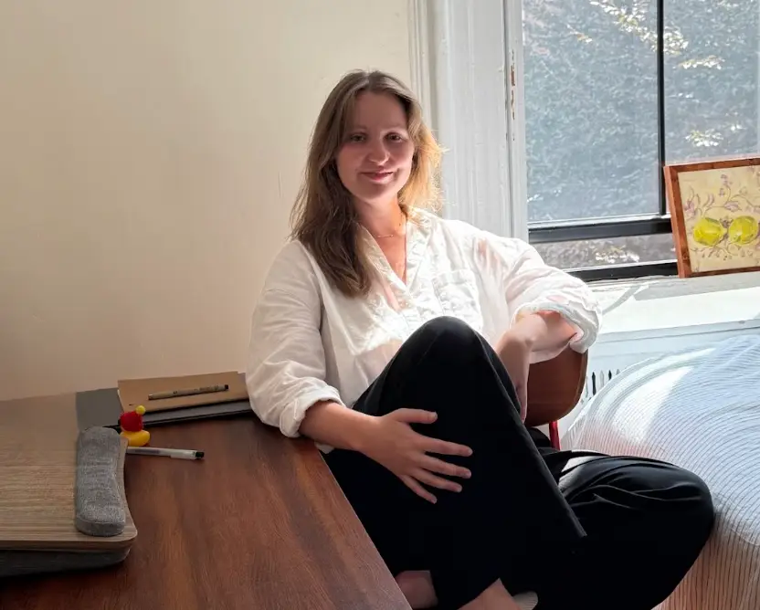 The writer wearing a white top and black pants, sitting at a wooden desk steps away from her bed.