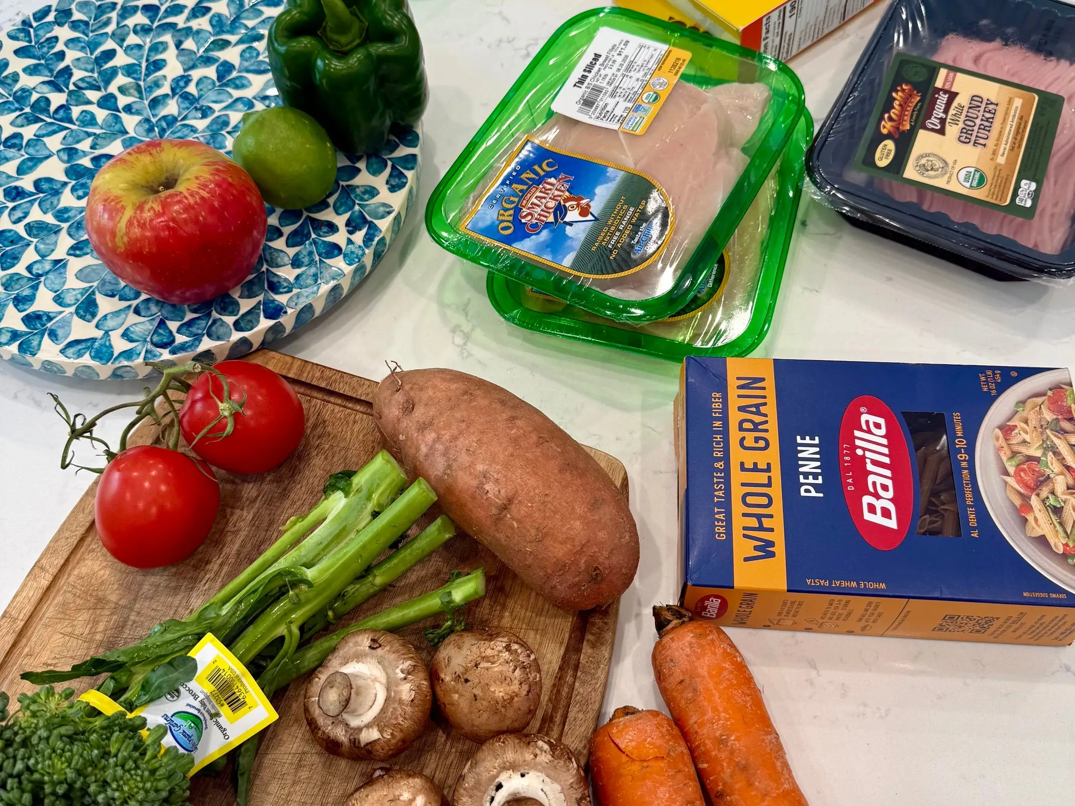 A spread of eggs, chicken, ground turkey, whole-grain penne, fruits, and vegetables on a kitchen table.