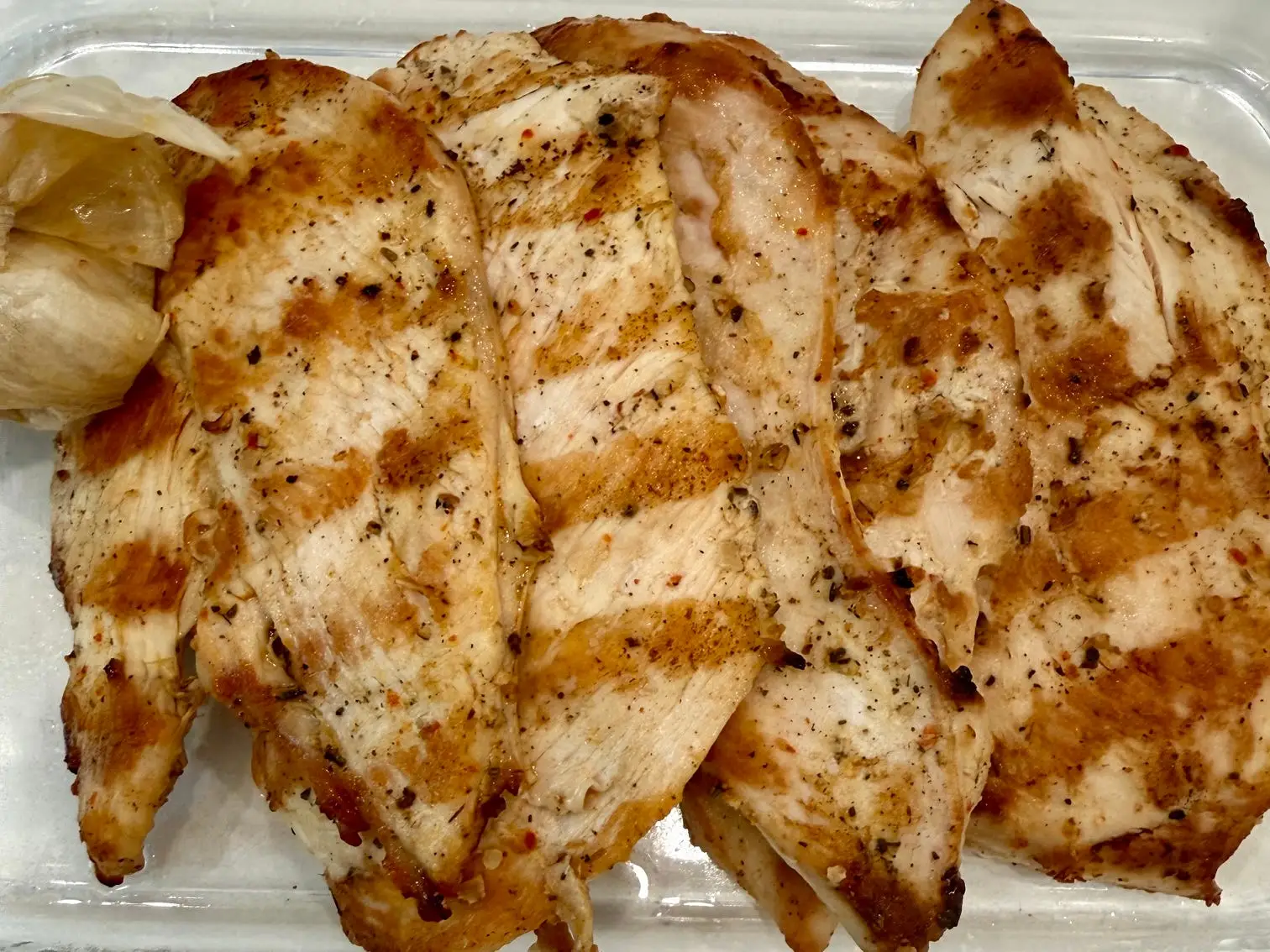 Sliced seasoned grilled chicken breast rests in a clear glass dish.