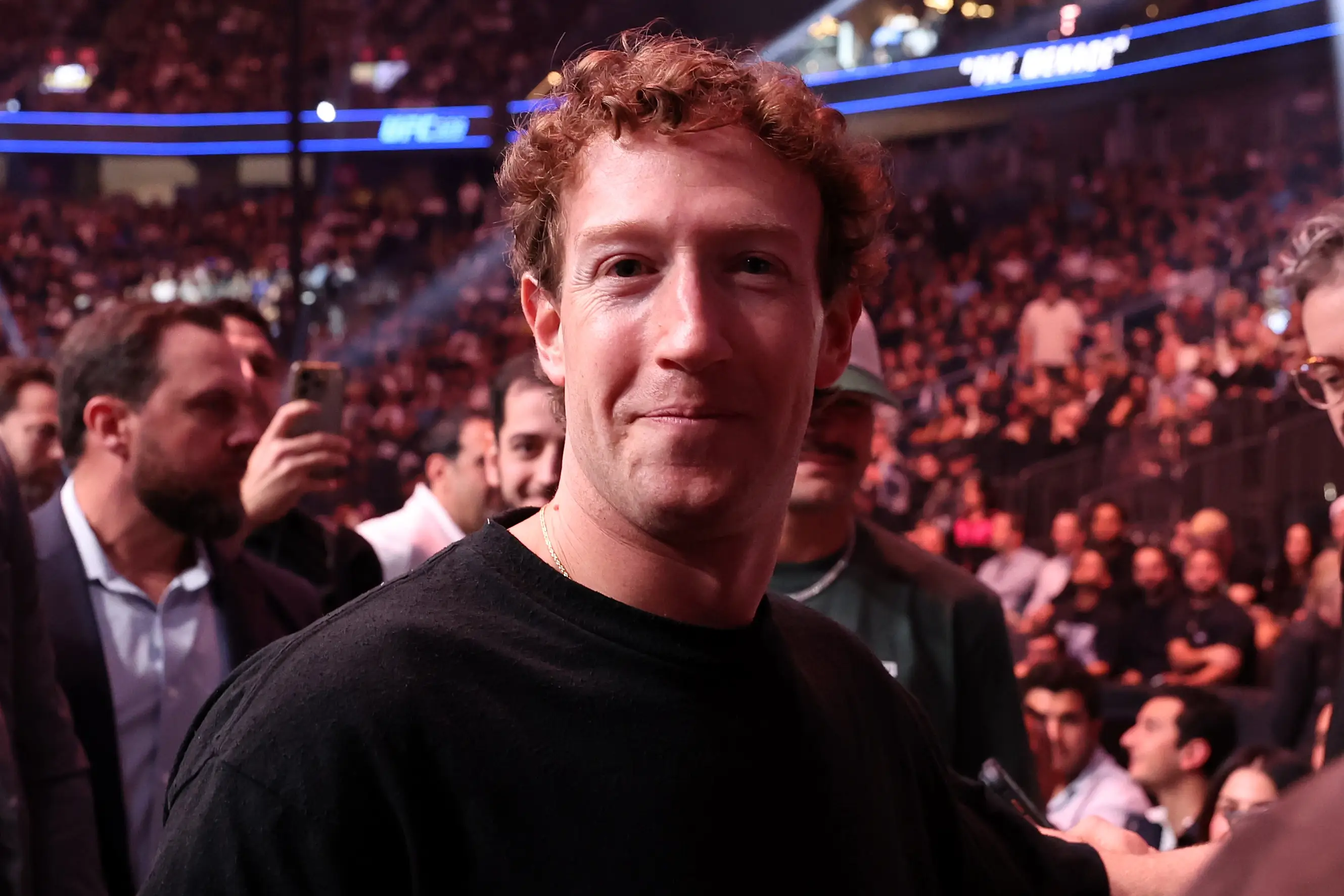 Mark Zuckerberg stands among spectators in a crowded indoor arena during a live event.
