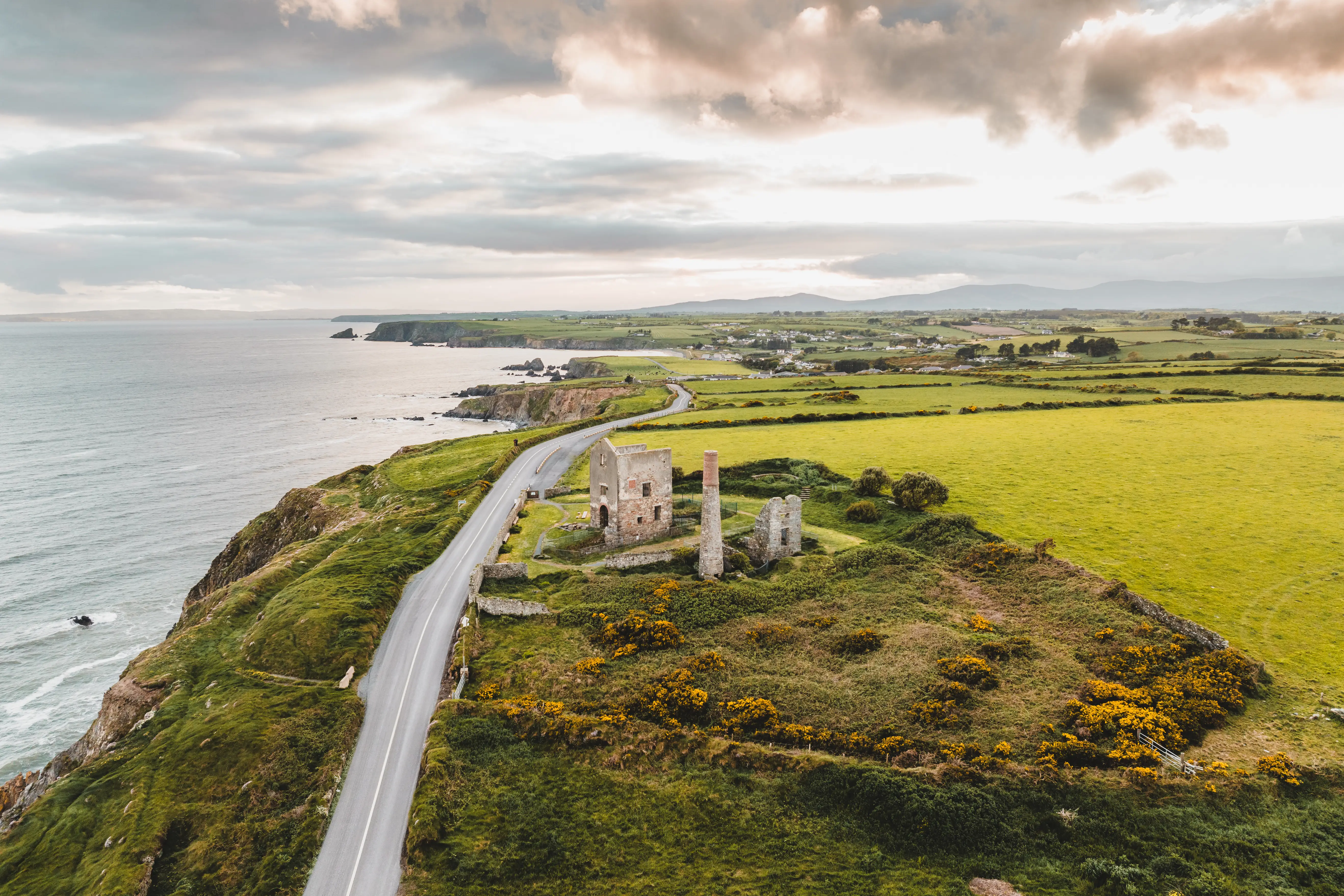 Coastal road passes stone ruins beside green fields and the sea under a cloudy sky.