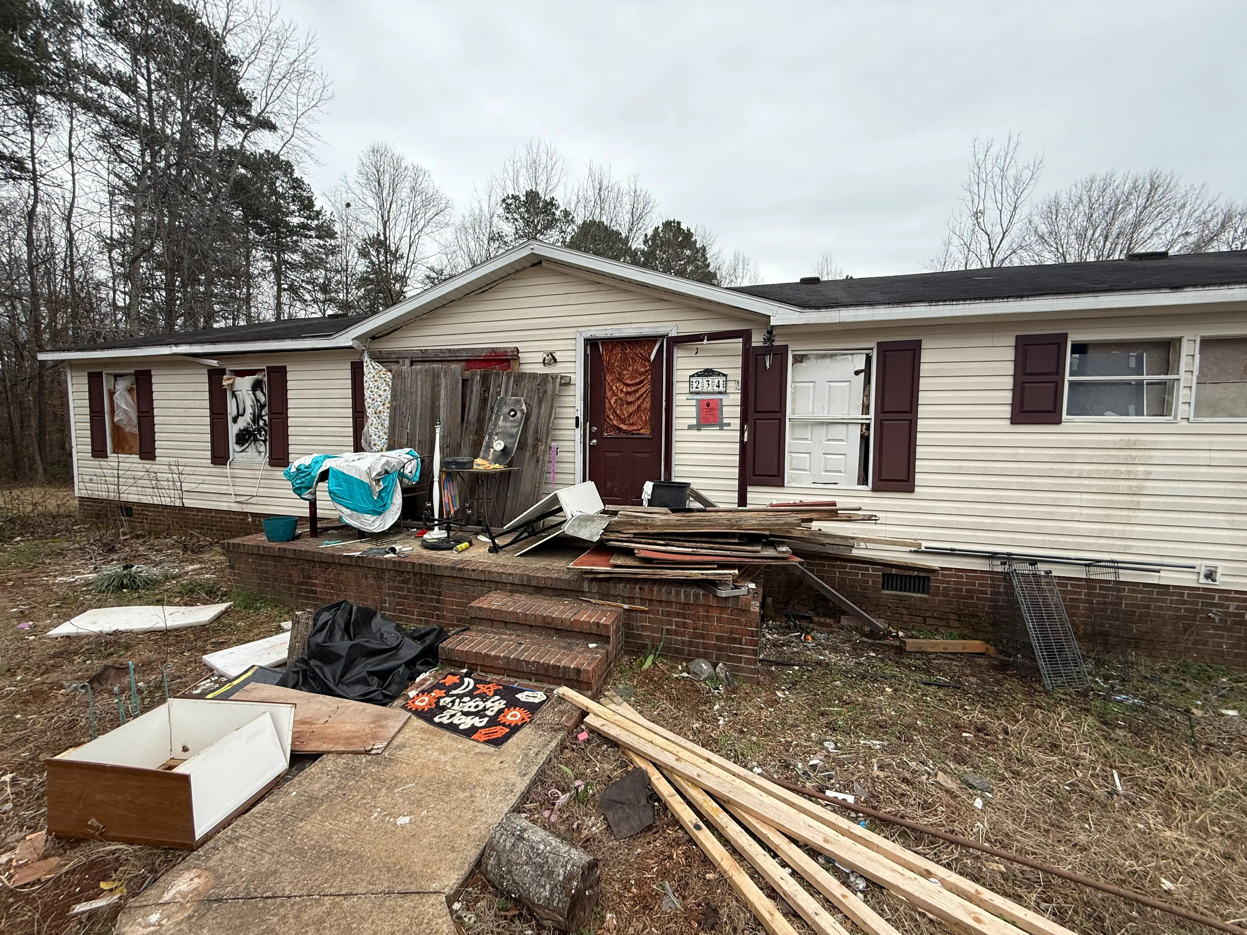 Dilapidated beige manufactured home with boarded openings and scattered debris across the front porch and yard.