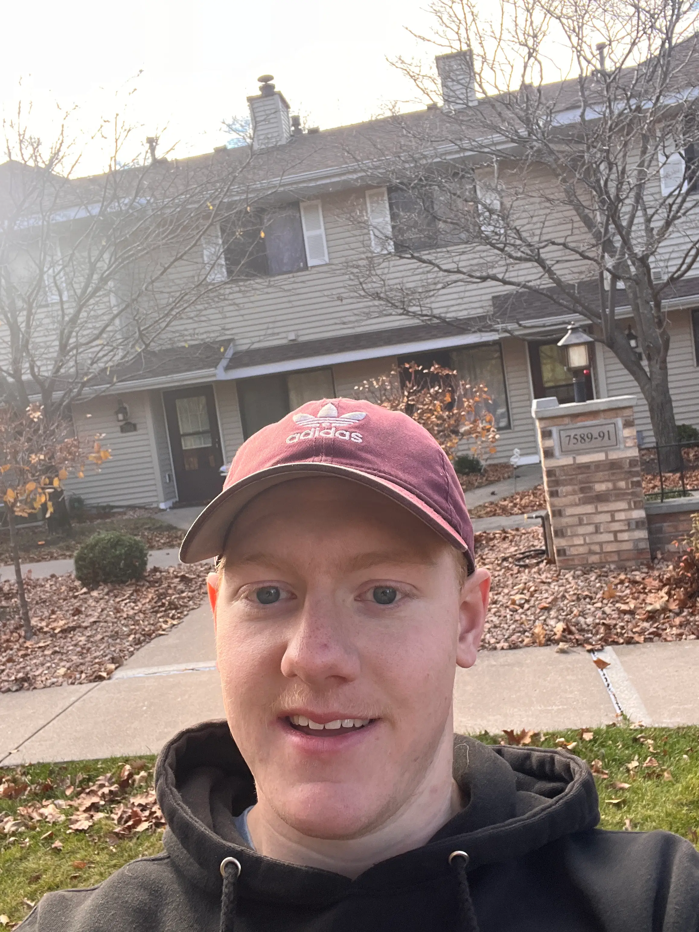 Person taking a selfie outside townhouse-style homes while wearing a burgundy cap.
