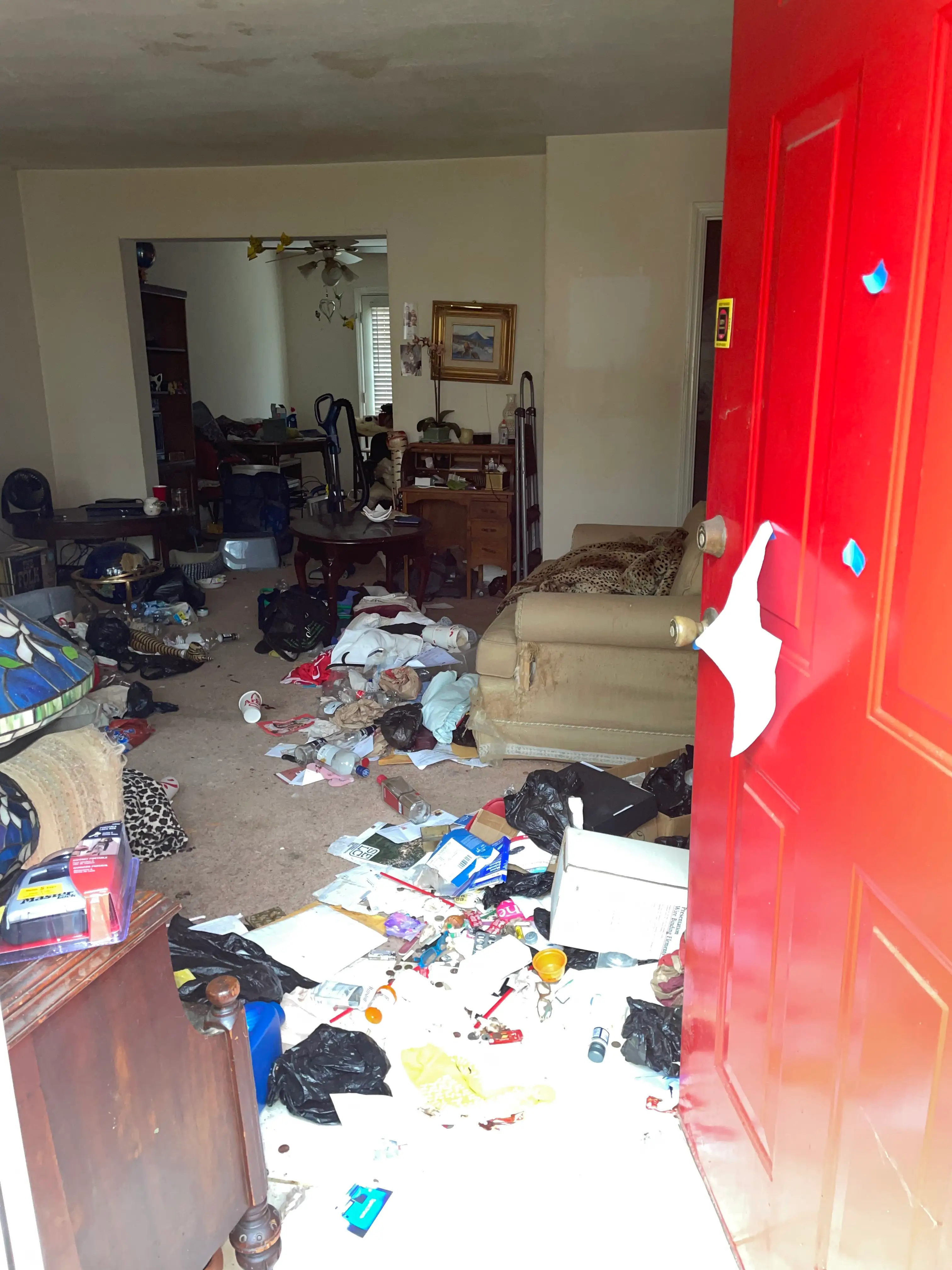 A red front door opens into a cluttered living room with scattered trash, furniture, boxes, and household items.