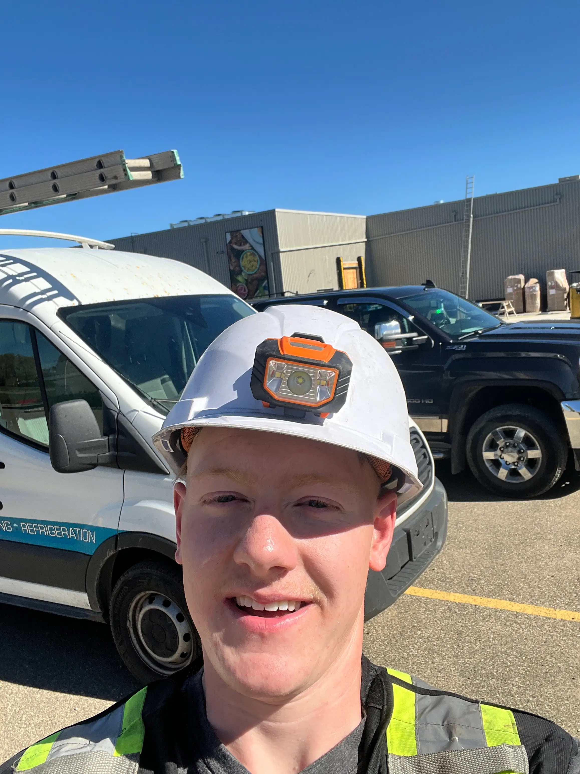 Worker wearing a white hard hat and high-visibility vest takes a selfie in an industrial parking lot.