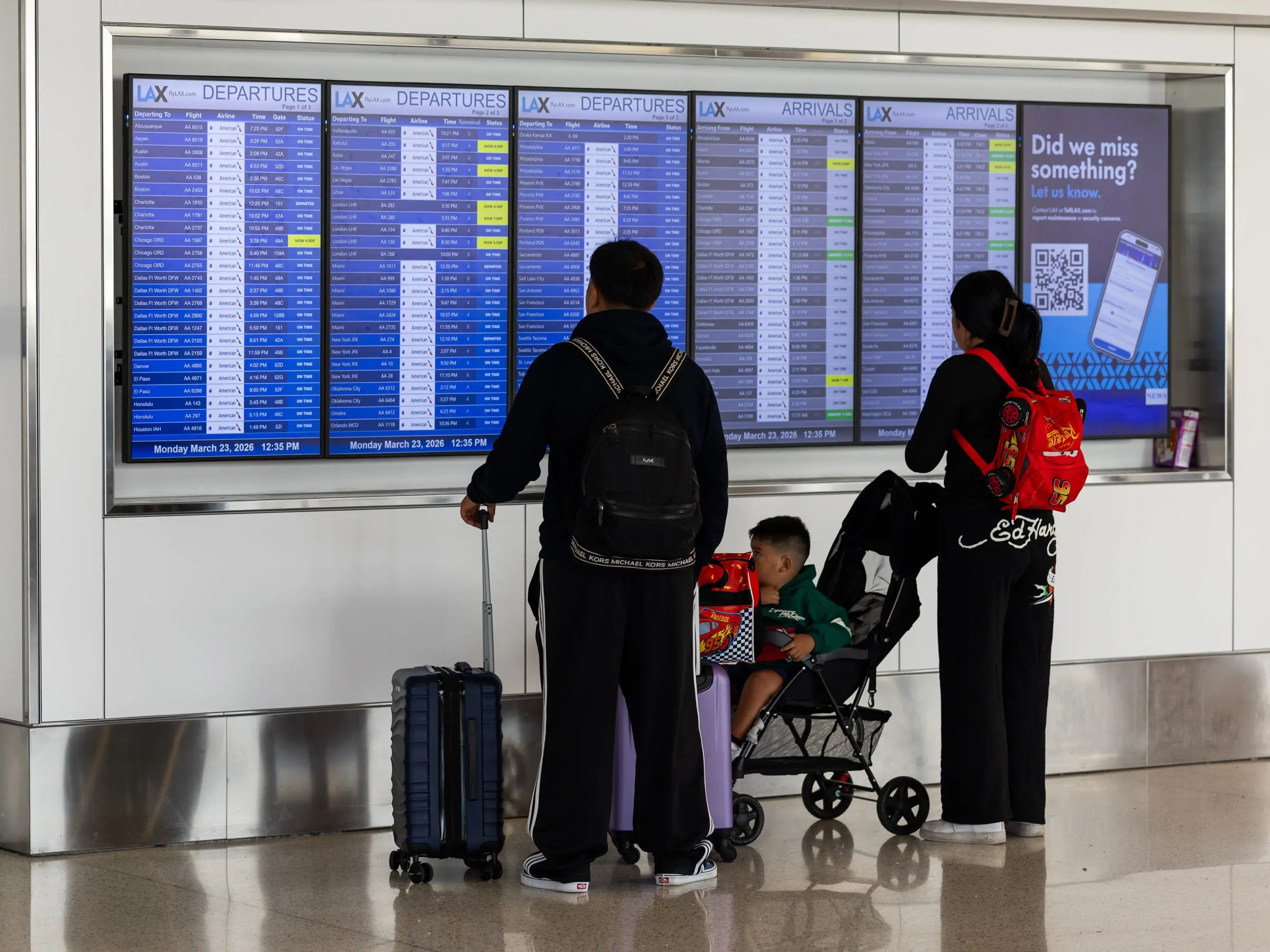 Travelers with luggage and a stroller stand in front of LAX departures and arrivals flight information screens.
