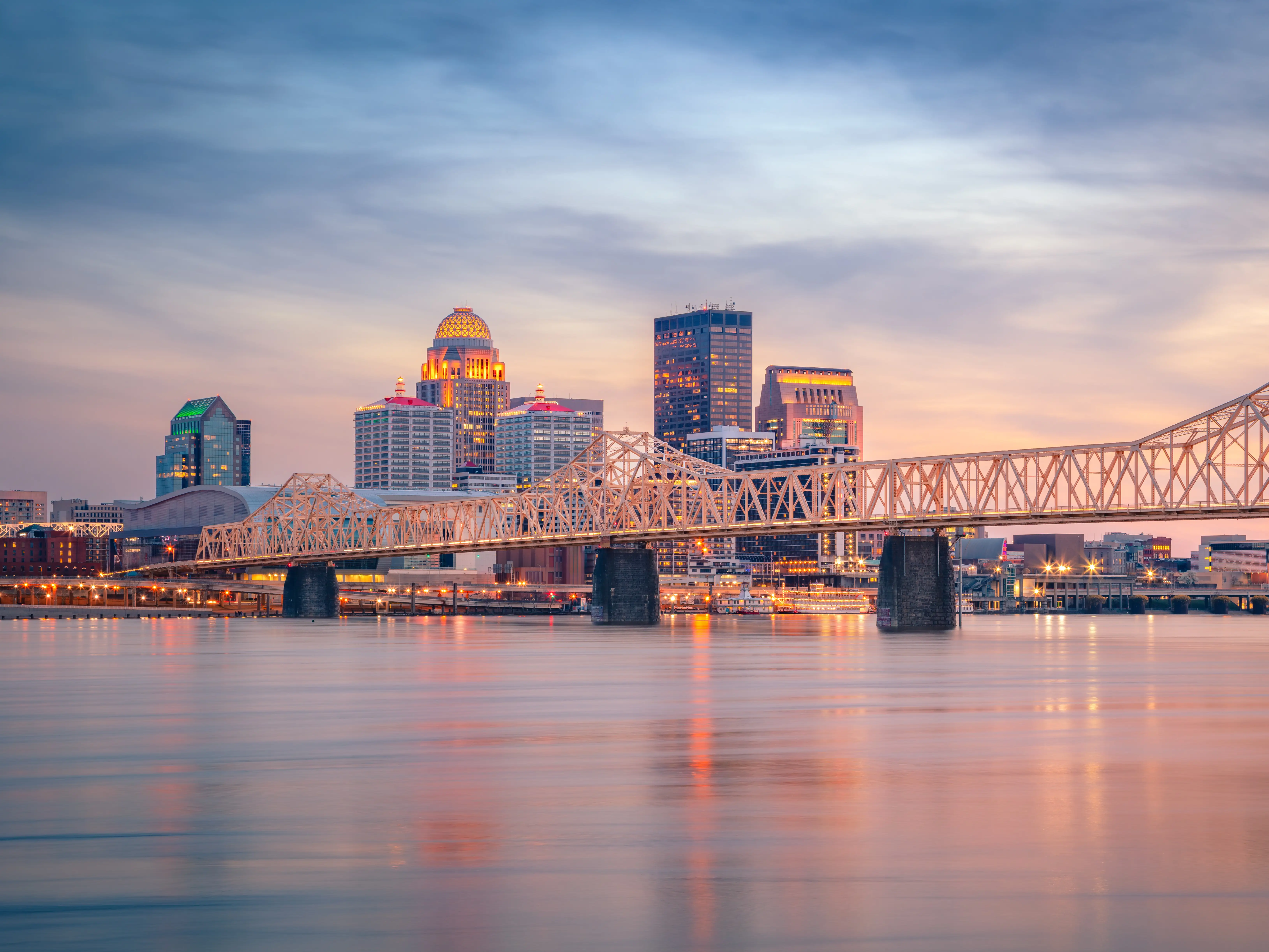 Louisville, Kentucky's downtown skyline is seen at sunset with buildings. The skyline reflects in the Ohio River, showcasing the water's calm surface. A prominent bridge, the John F. Kennedy Memorial Bridge, extends across the river, featuring intricate truss work. The sky is painted with soft hues of blue, pink, and orange, creating a serene atmosphere typical of spring.