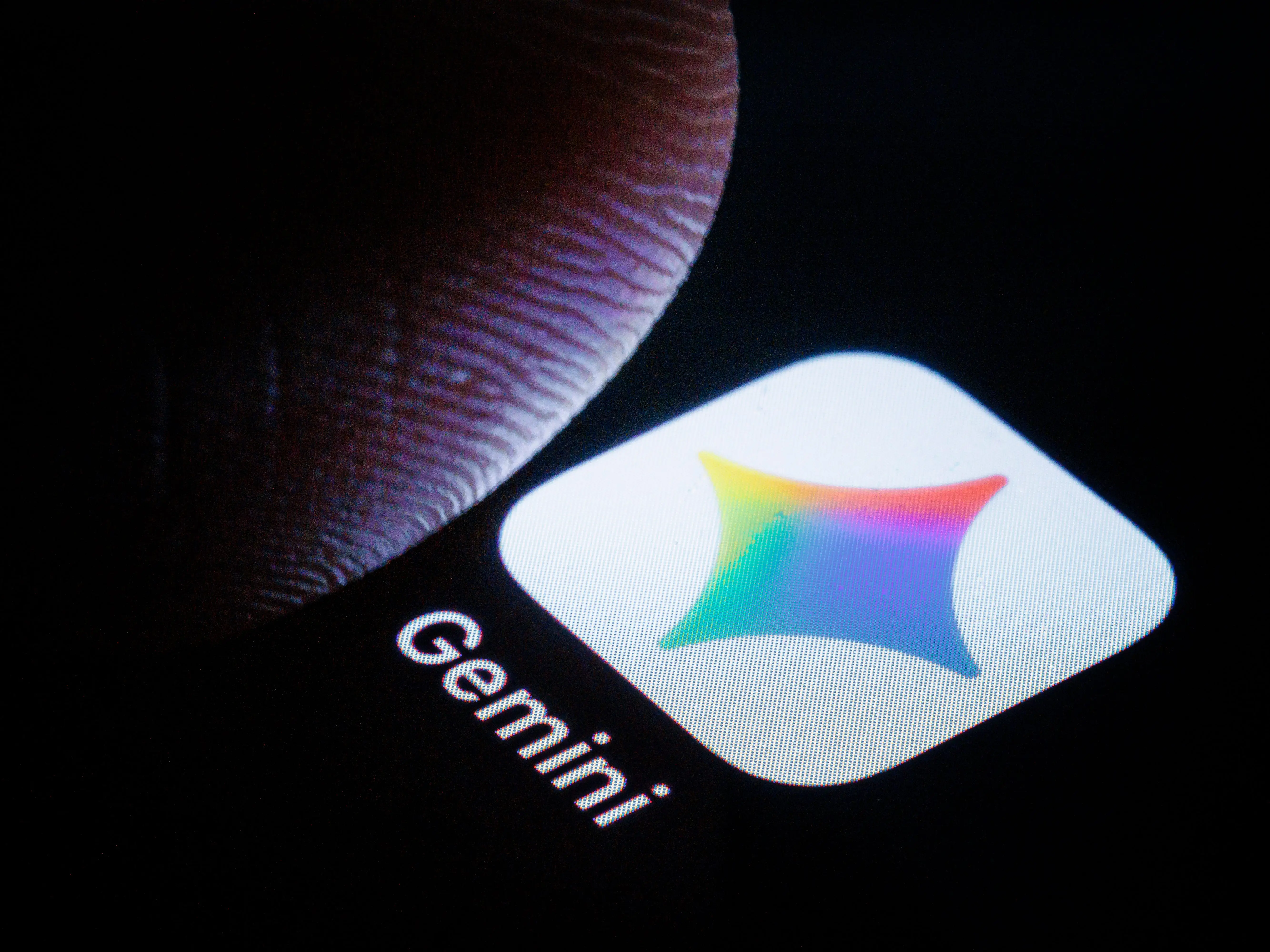Close-up smartphone screen shows the Gemini app icon and label beside a fingertip in low light.