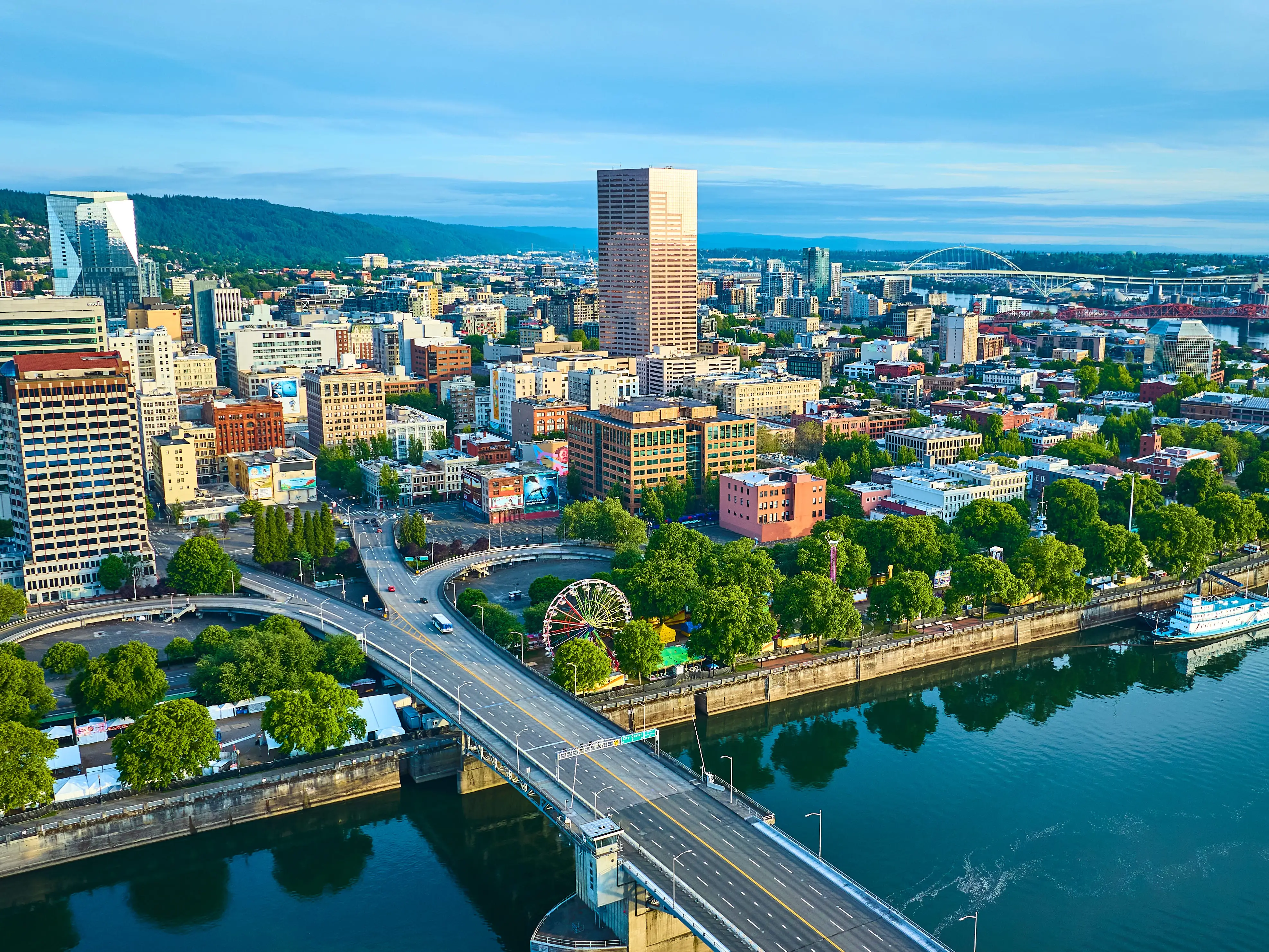 Aerial view of Portland, Oregon, showcasing the vibrant downtown with a mix of modern and classic skyscrapers. The Willamette River with a bridge and paddlewheel riverboat adds scenic beauty and historical charm.