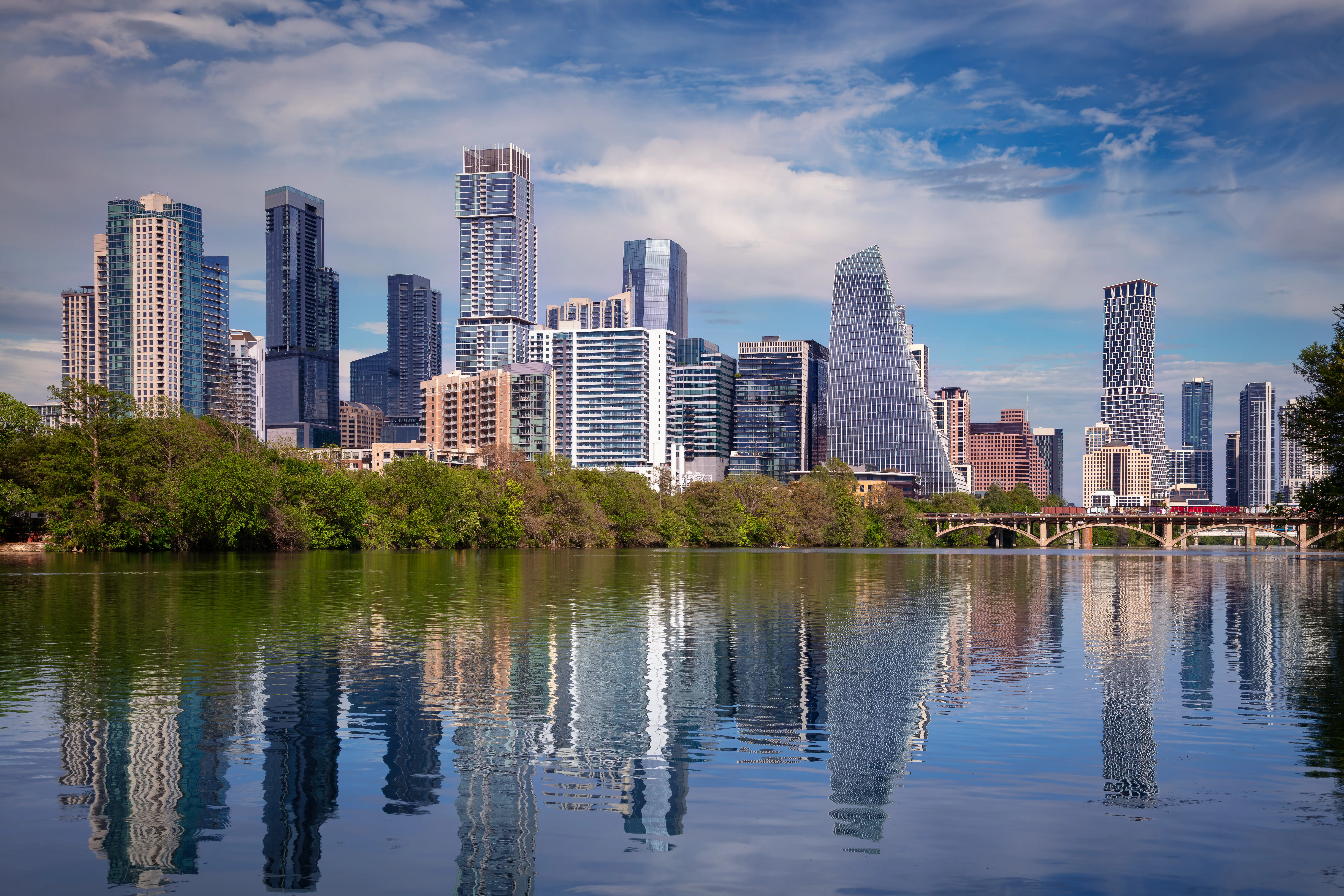 Austin, Texas skyline with modern skyscrapers and unique architectural designs against a blue sky. The foreground features a calm body of water reflecting the buildings and surrounding green trees. The scene captures a mix of urban architecture and natural beauty, showcasing Austin's vibrant cityscape and lush environment.