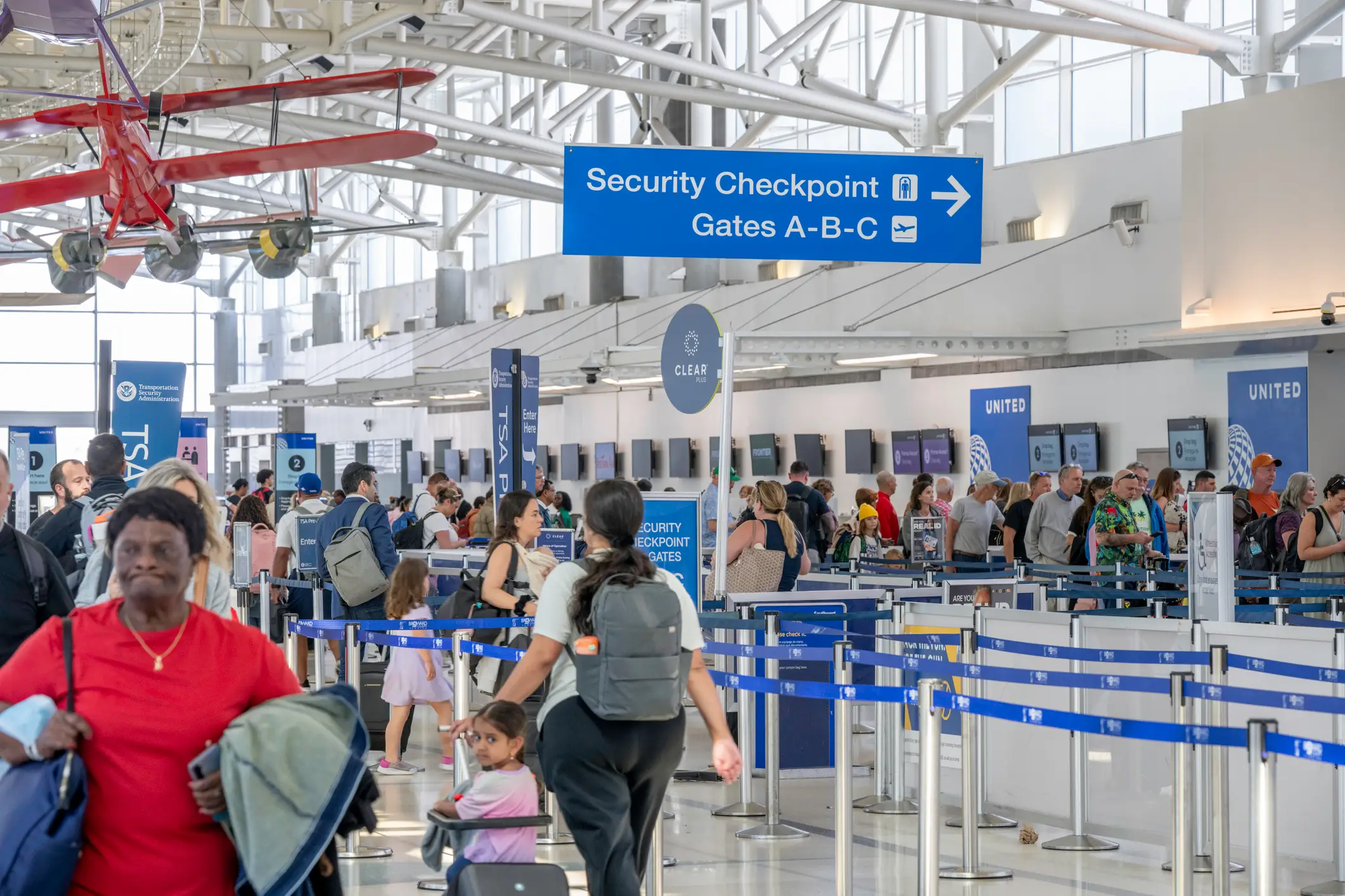 Travelers queue at an airport security checkpoint below signs for Gates A-B-C and United counters.