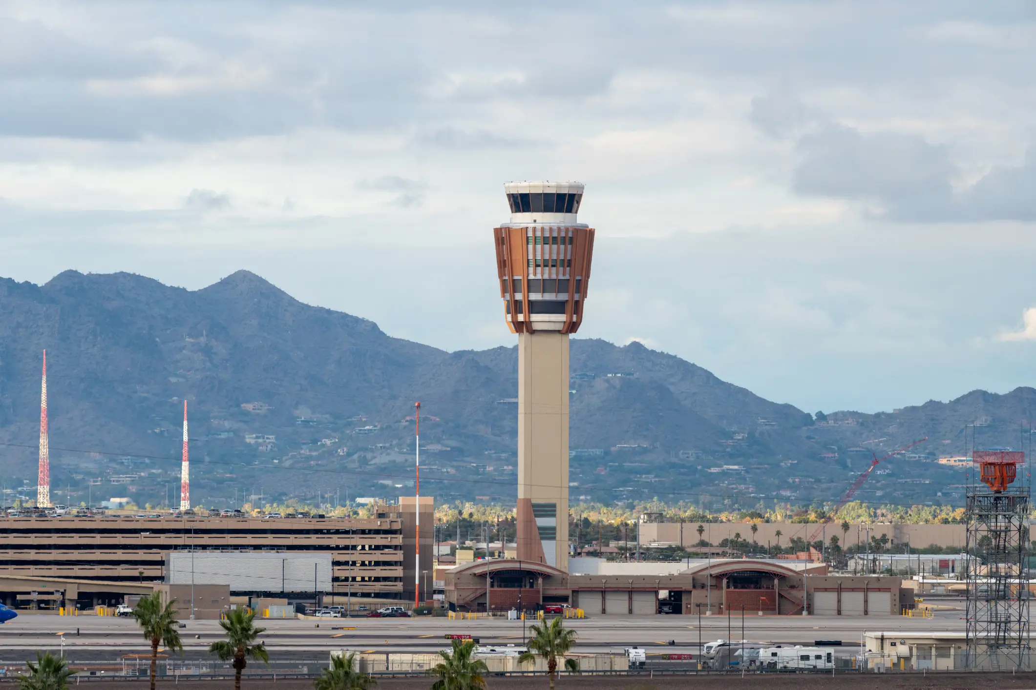 Airport control tower and terminal buildings stand before mountains under a cloudy sky.