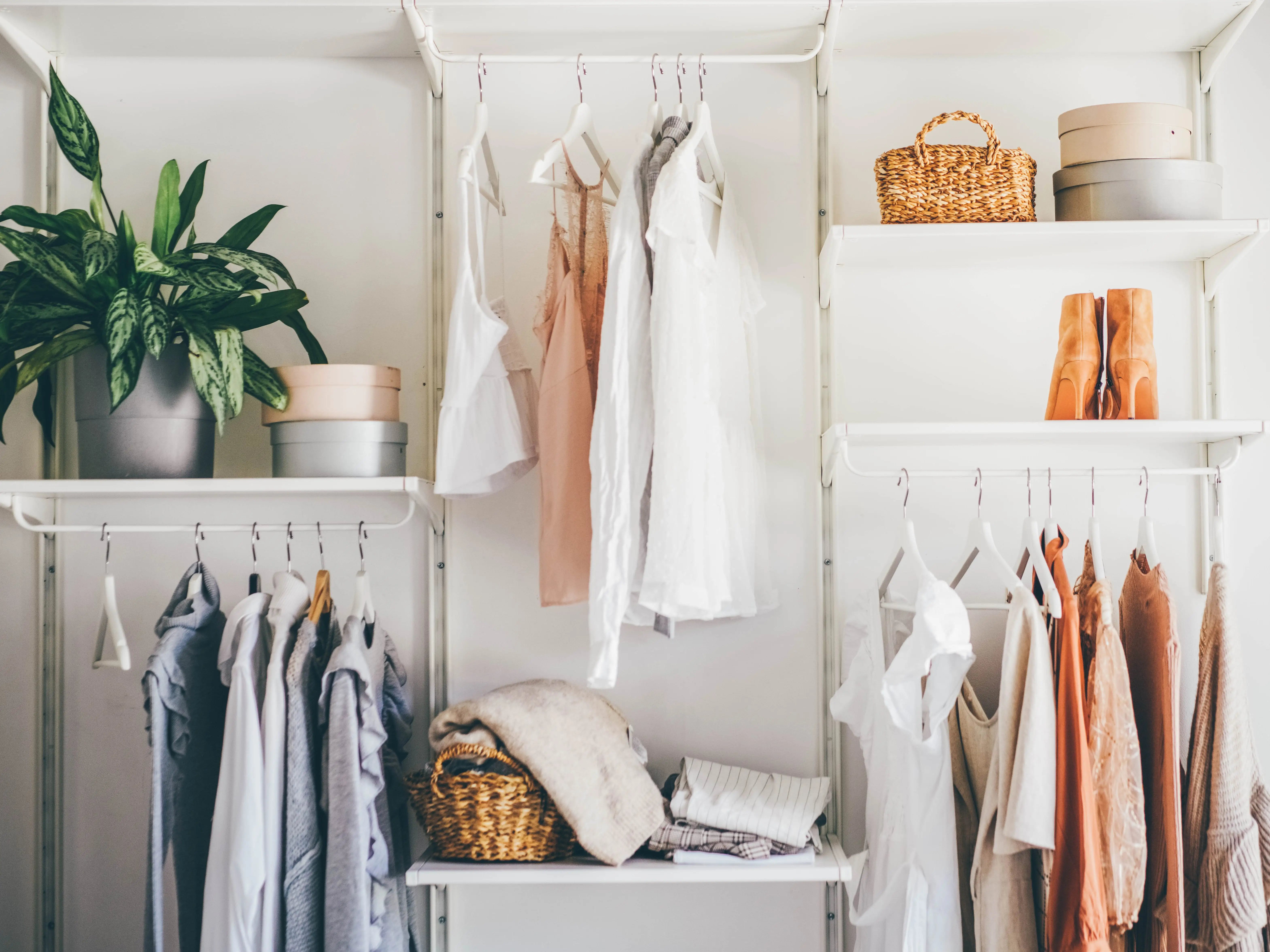 Open white closet shelves display hanging clothes, woven baskets, boots, storage boxes, and a potted plant in an organized fashion.