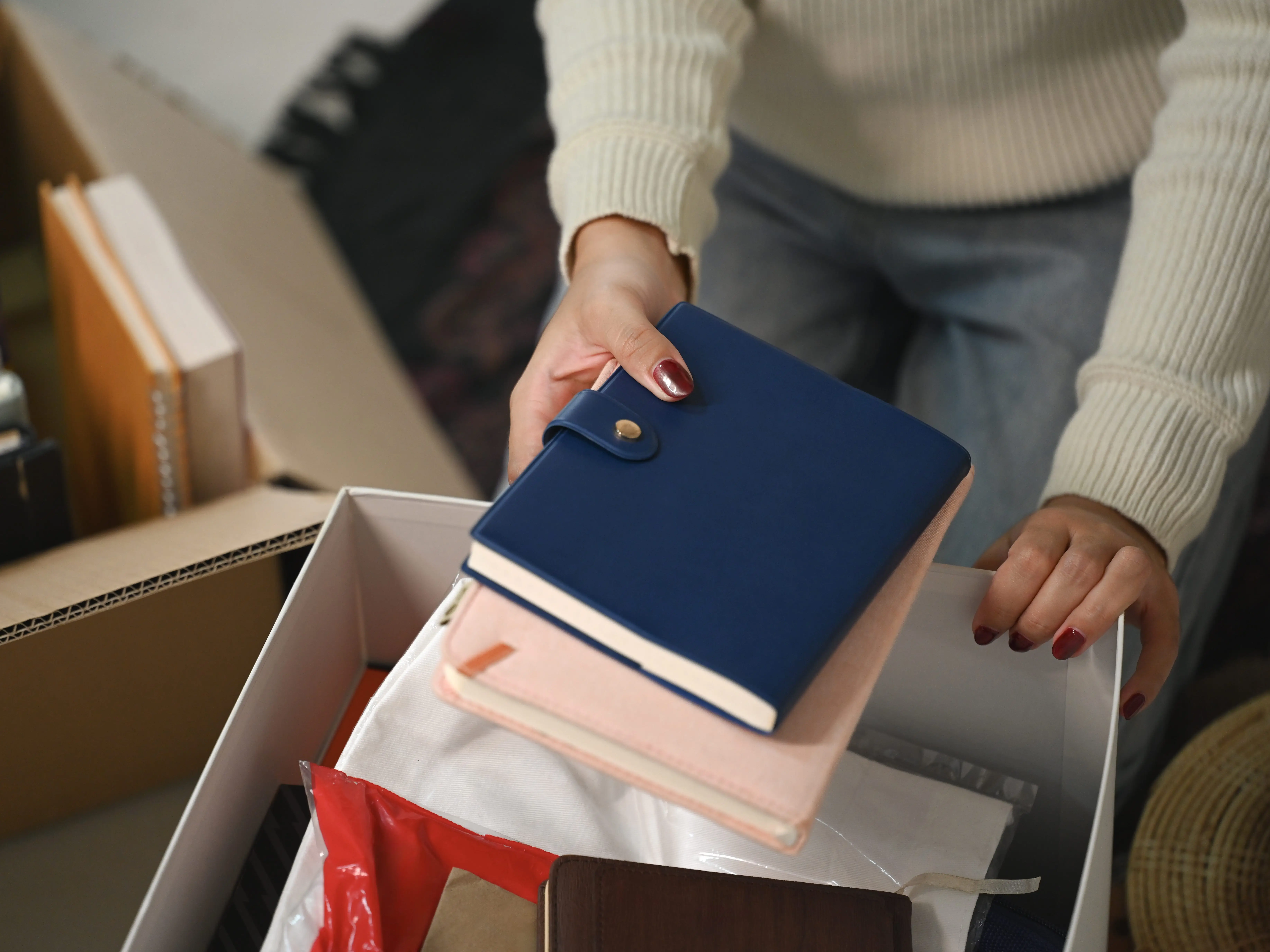 Hands place a blue notebook into a storage box filled with books, folders, and fabric items.