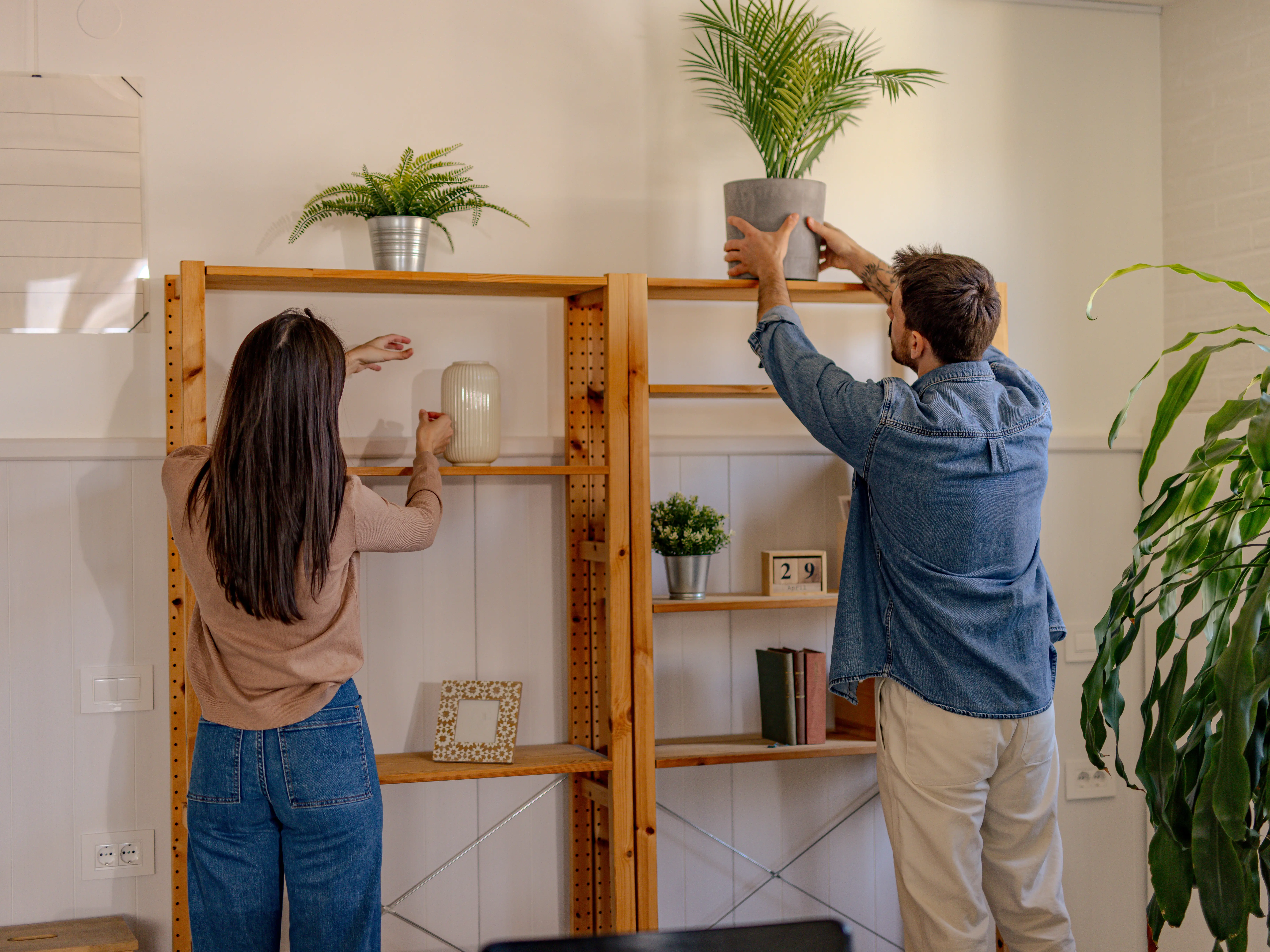Two people arrange potted plants and decor on wooden shelving in a bright indoor room.