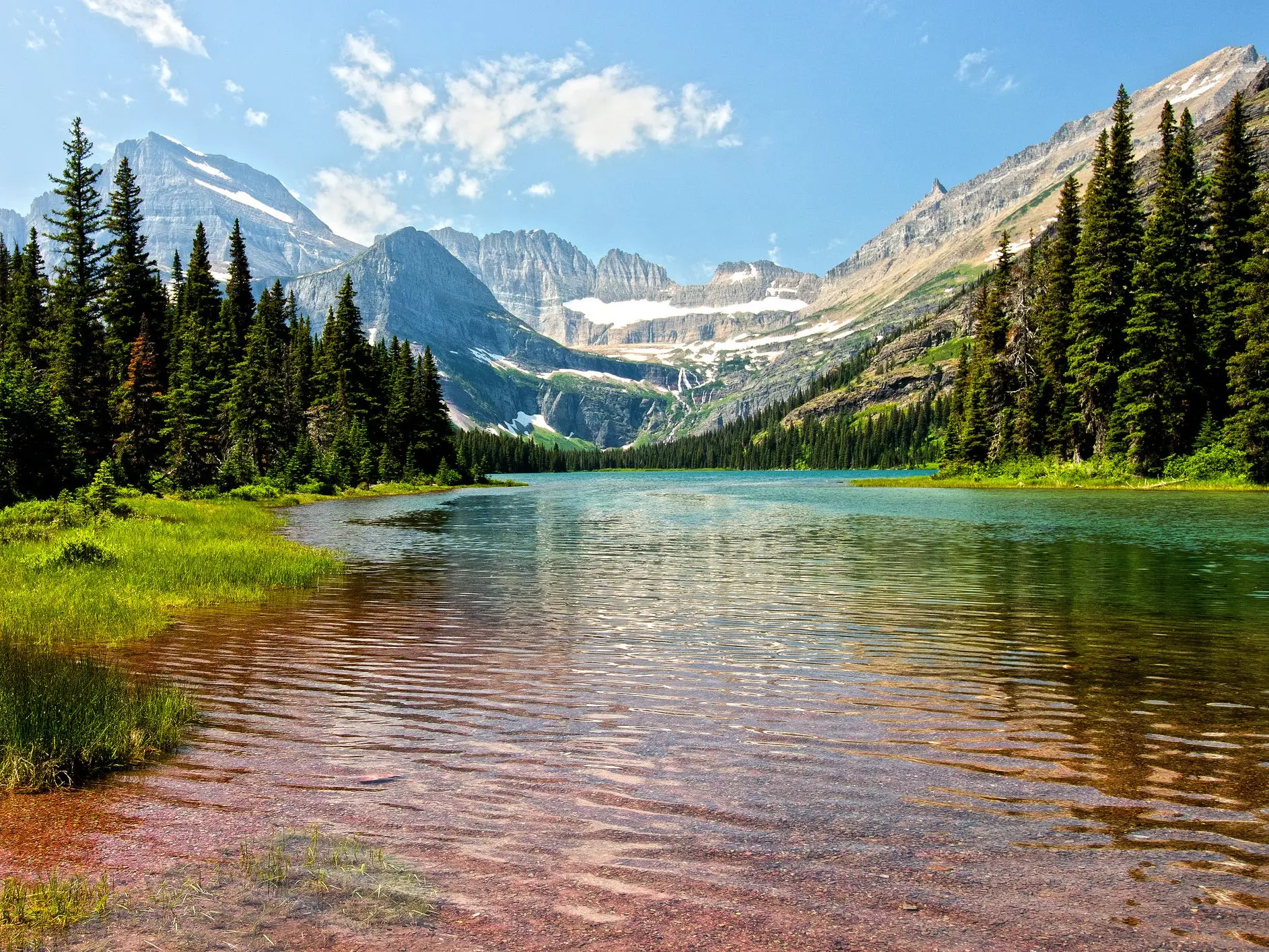 Clear alpine lake in Glacier National Park bordered by evergreen forest with rugged snow-dusted mountains under a blue sky.