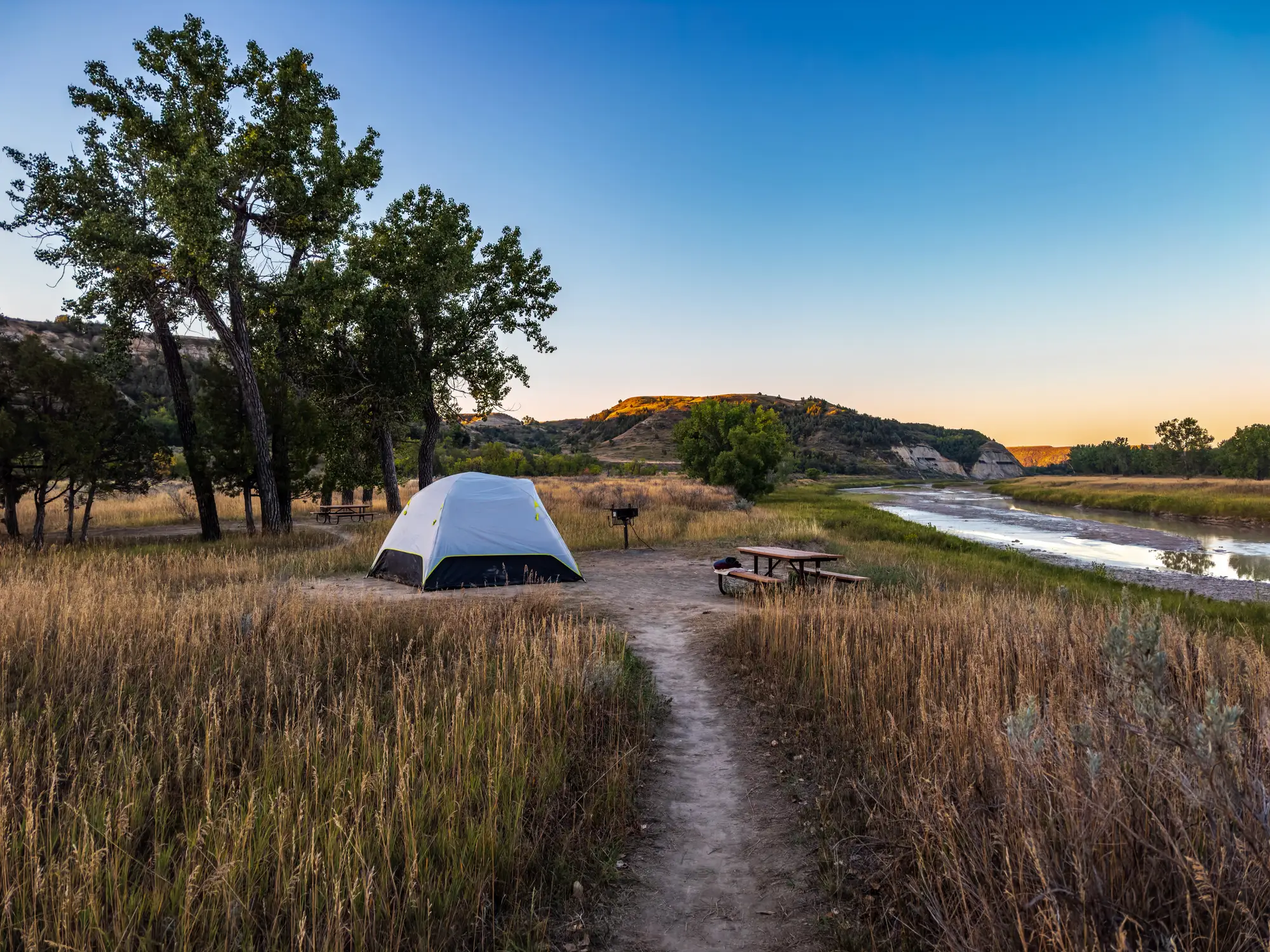 Riverside campsite at Theodore Roosevelt National Park in North Dakota with a white tent, picnic table, trees, tall grass, and sunlit hills at dusk.