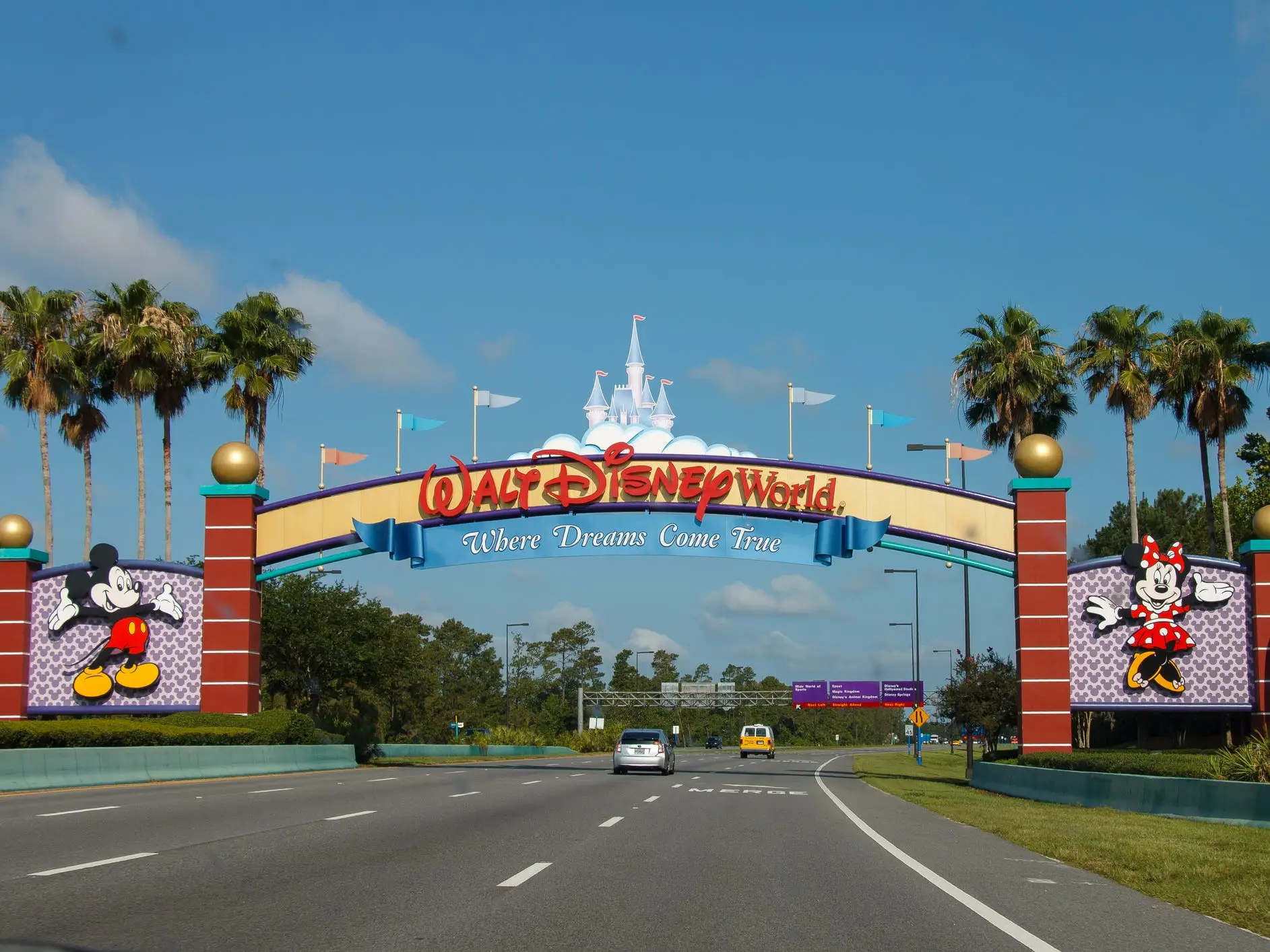 Walt Disney World entrance arch spans a roadway with Mickey Mouse and Minnie Mouse signs under a blue sky.