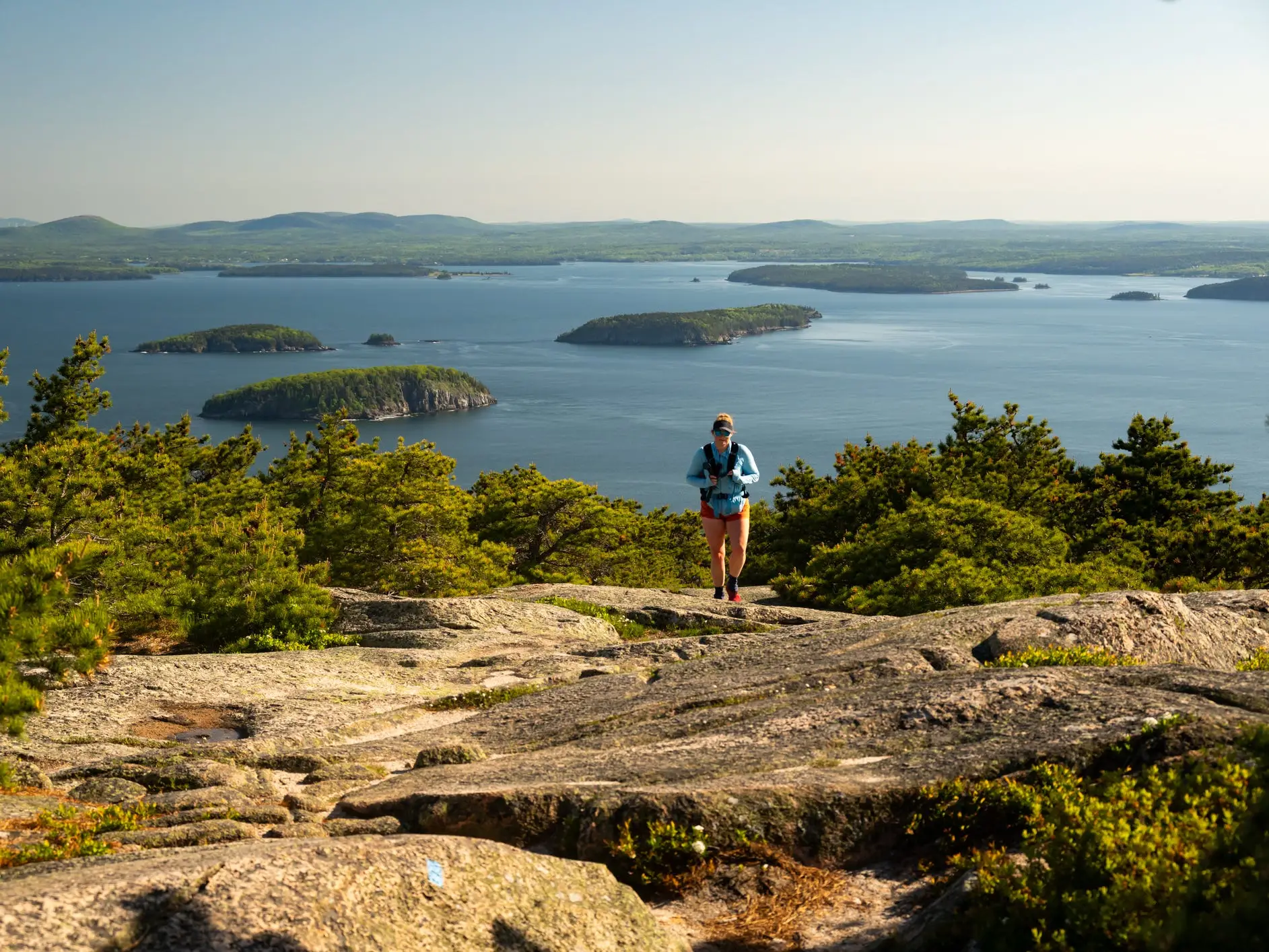 Hiker on a rocky coastal overlook in Acadia National Park with forested islands, blue water, pine trees, and distant hills.