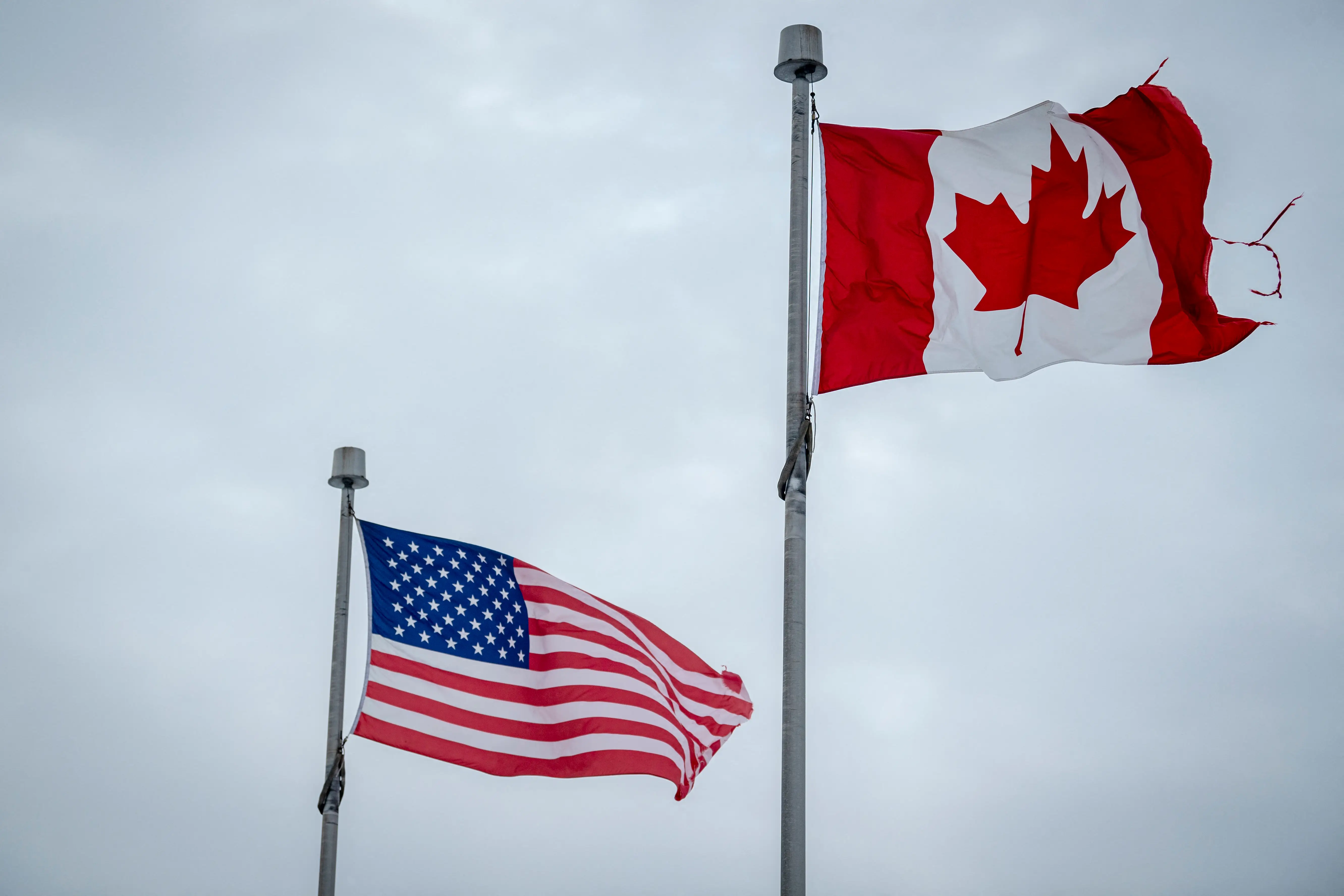 Tattered United States and Canada flags fly on separate poles against a cloudy gray sky.
