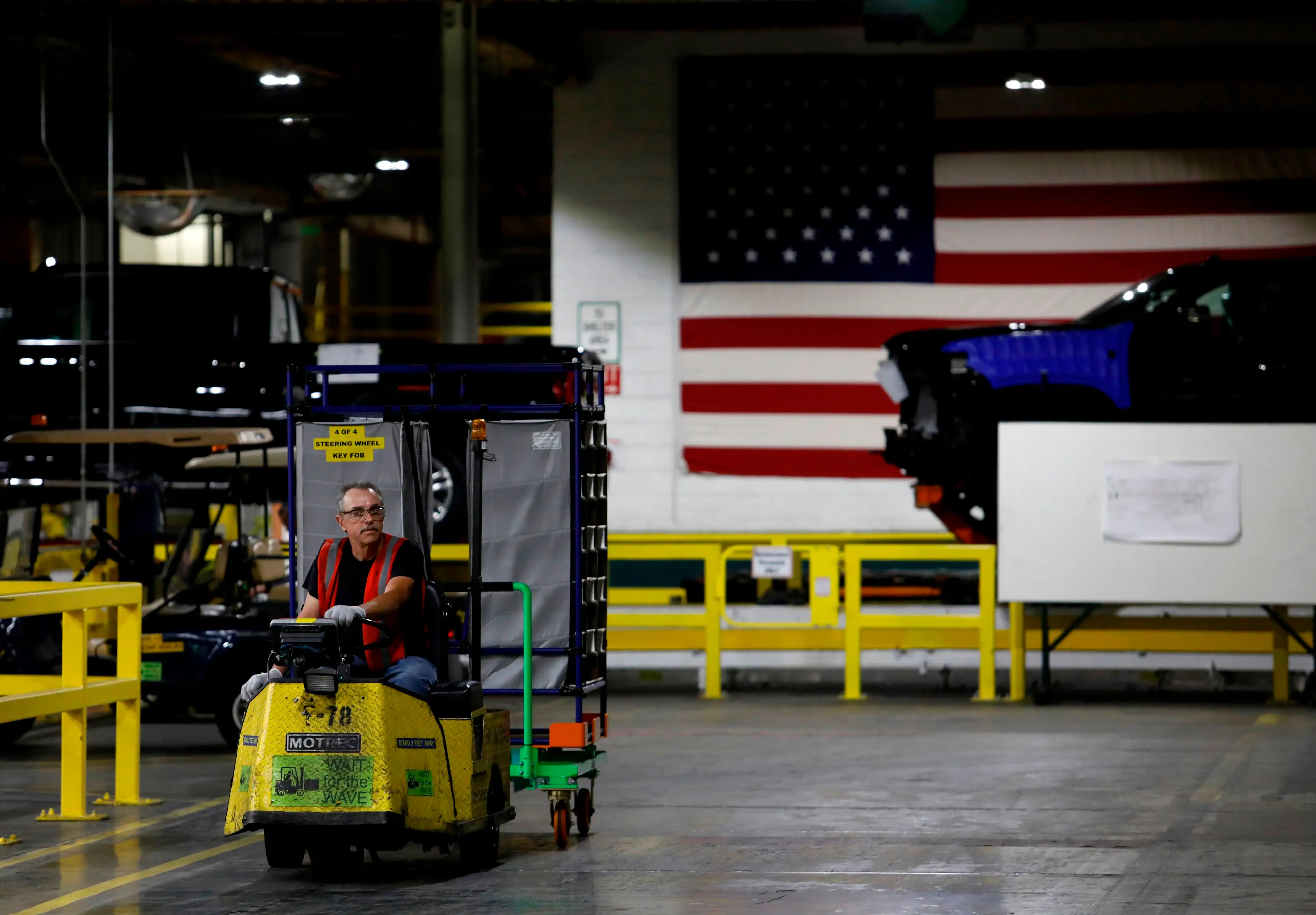 A GM employee drives a cart in an assembly plant. There's a large American flag behind a full-size car in the background.