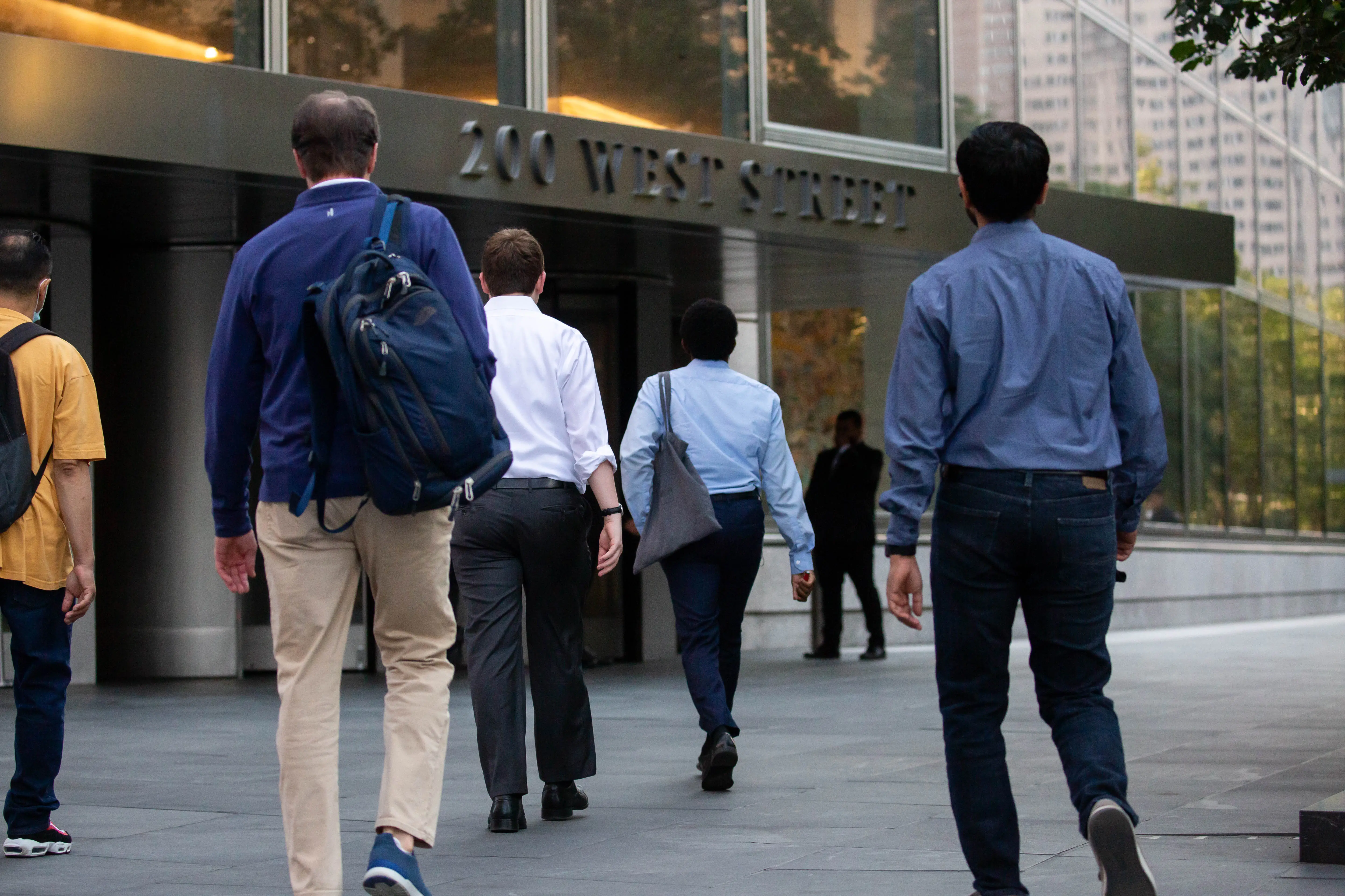 Pedestrians with backpacks walk toward a glass office building entrance marked “200 WEST STREET.”