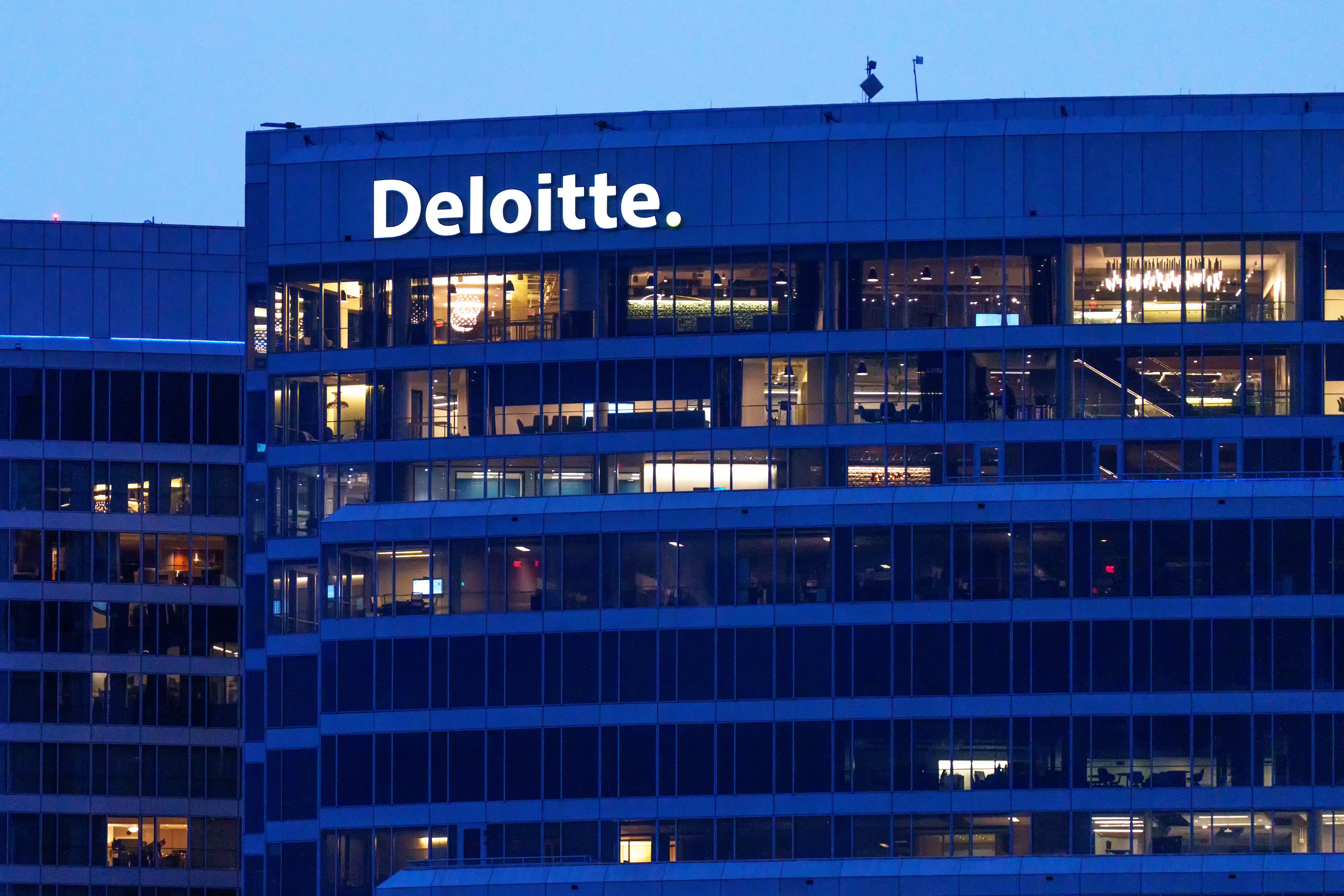 Deloitte sign illuminated on the upper floors of a glass office building at dusk.