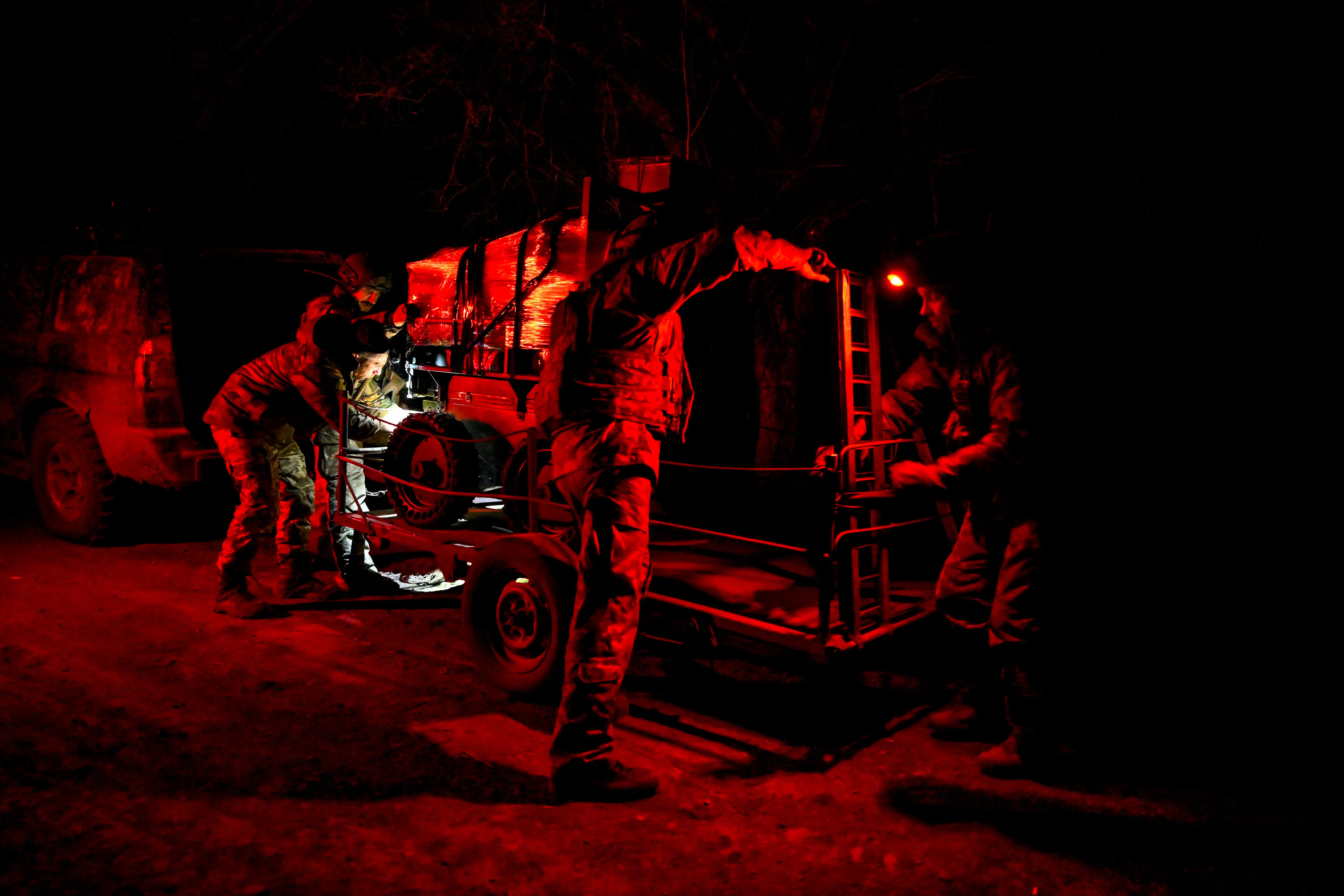 Uniformed personnel work around a trailer and vehicle under red light during a nighttime operation.