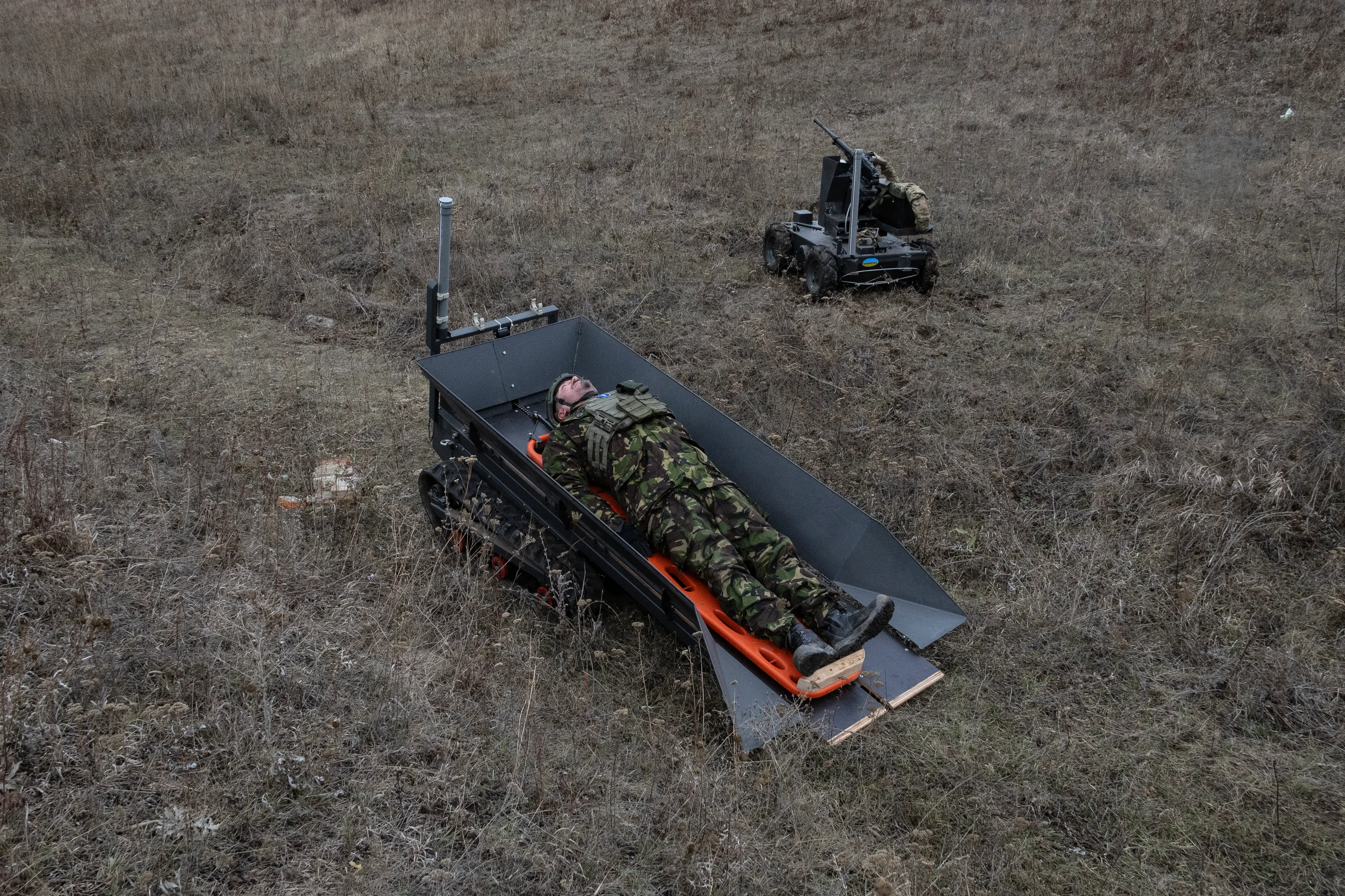 Person in camouflage lies on an orange stretcher atop a tracked robotic vehicle in a dry field.
