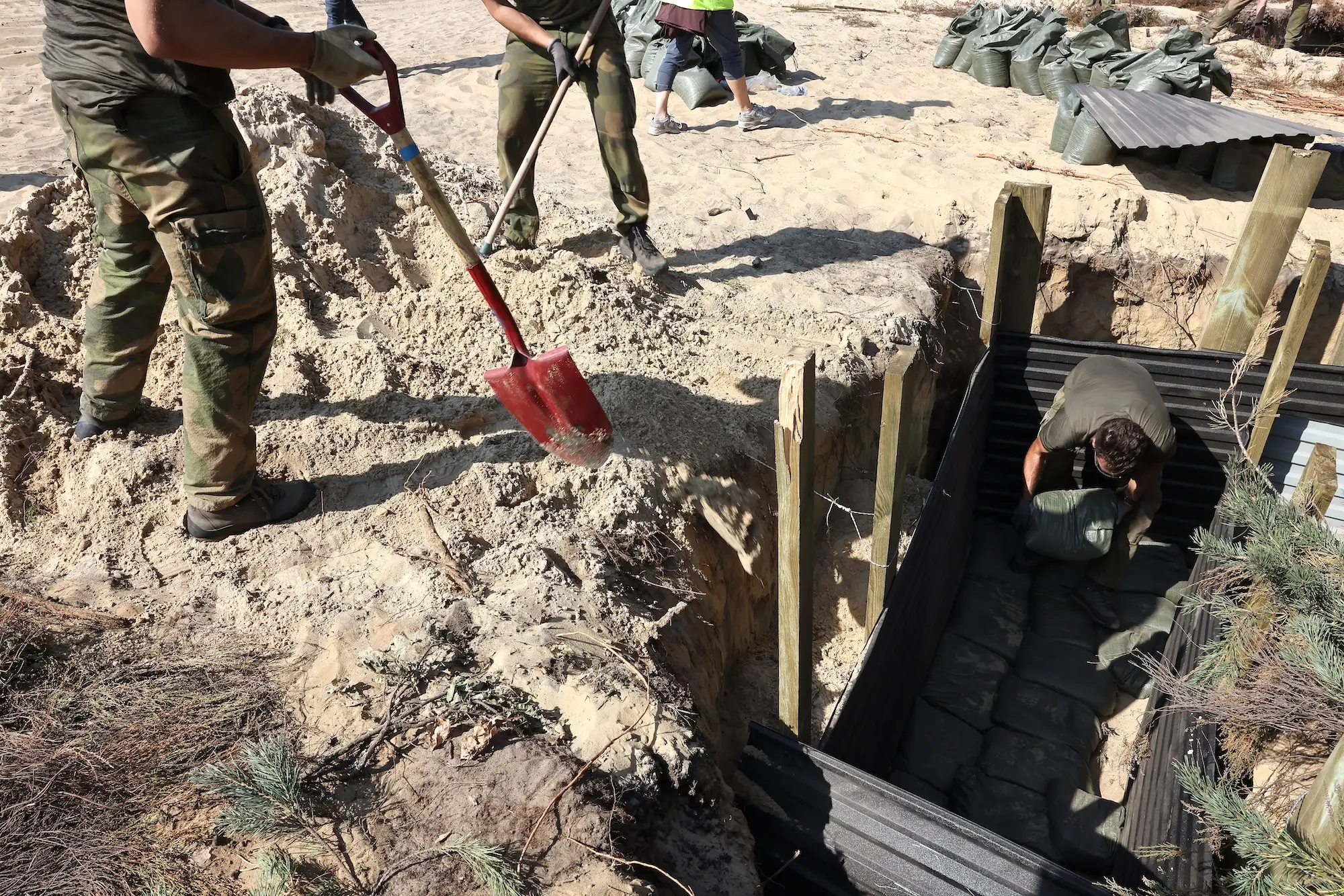 People in camouflage clothing shovel sand around a timber-lined trench filled with sandbags at a sandy outdoor site.