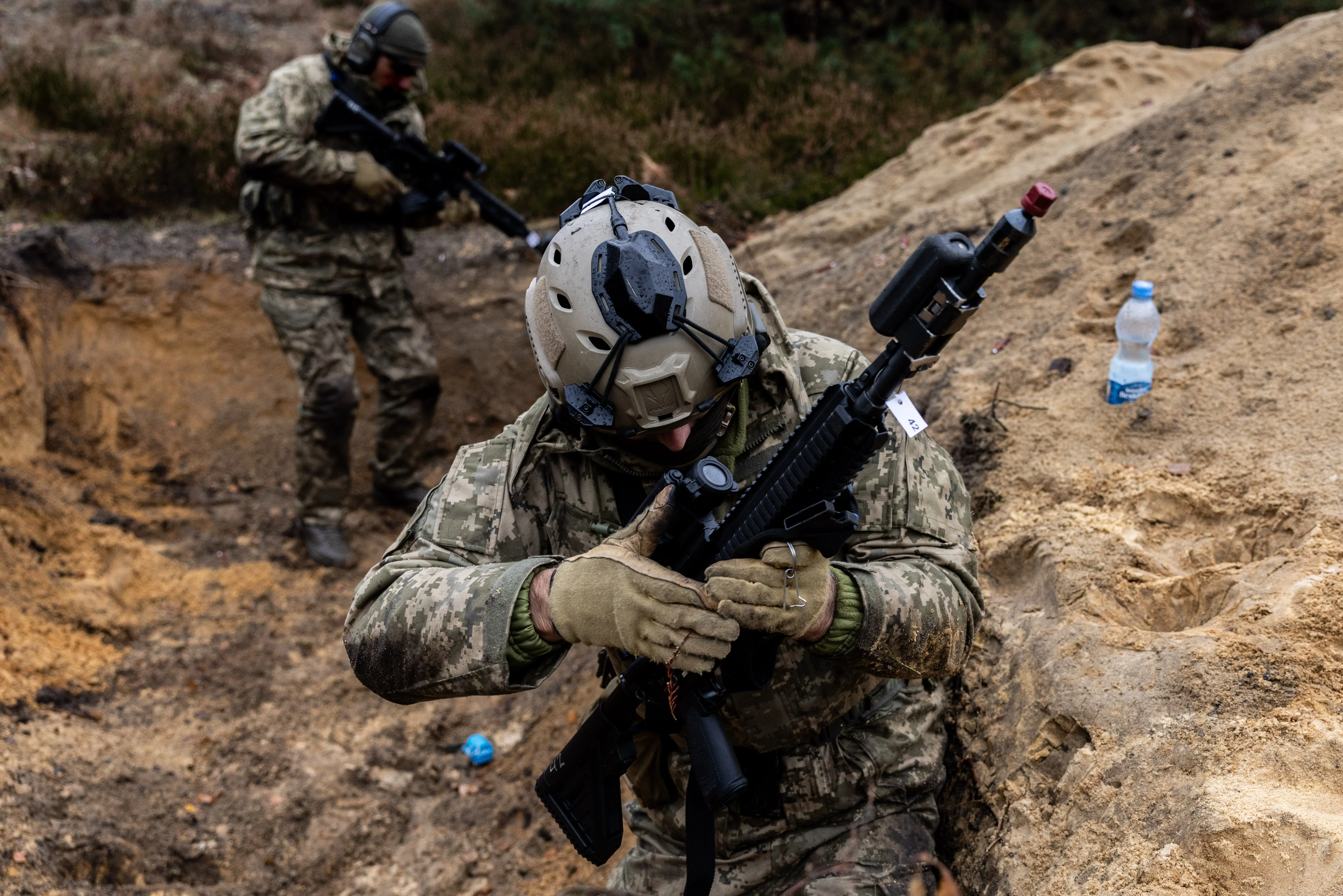Two camouflaged armed personnel move through a sandy trench with rifles during an outdoor military exercise.