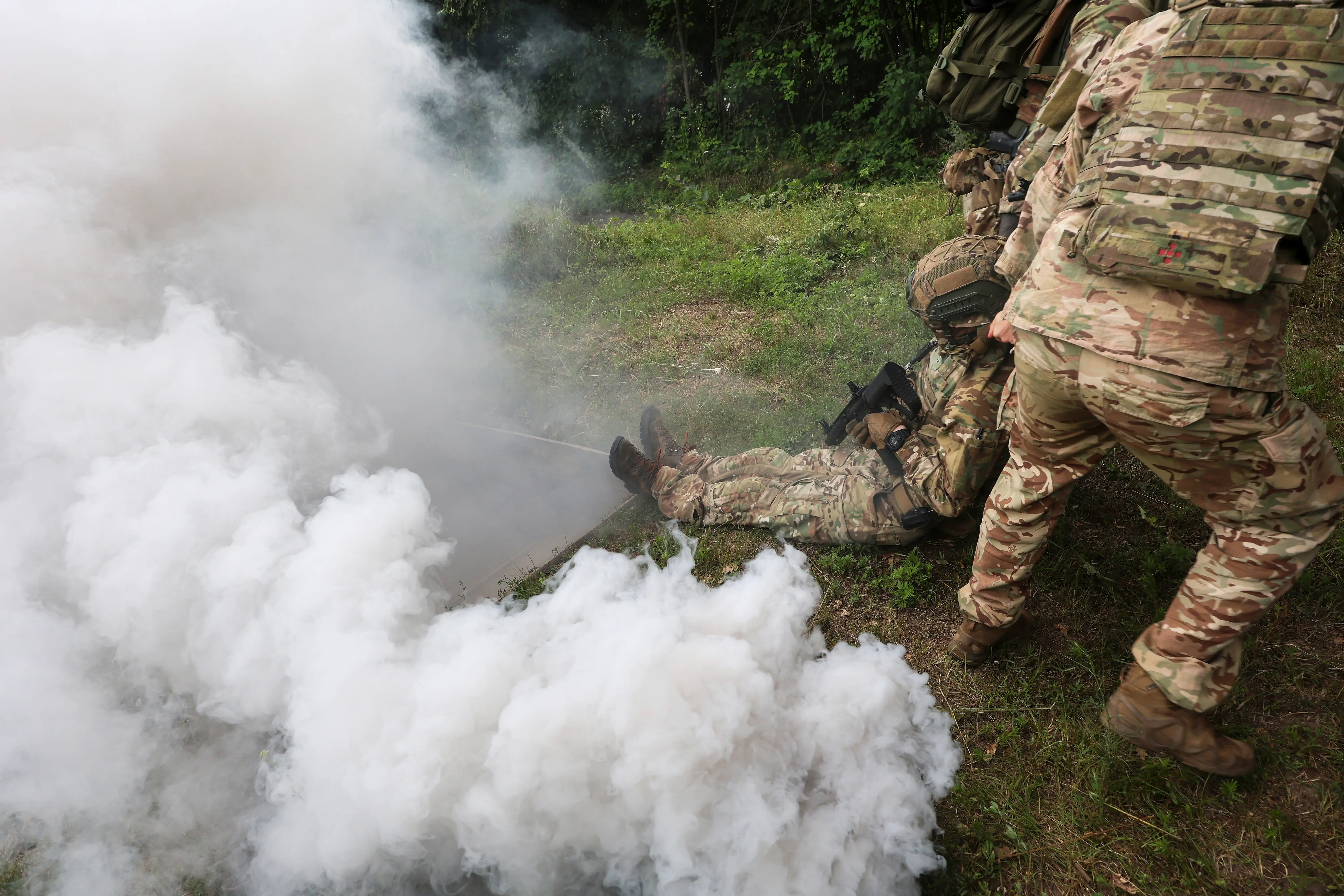 Camouflaged personnel with rifles move through thick white smoke in a wooded outdoor setting.