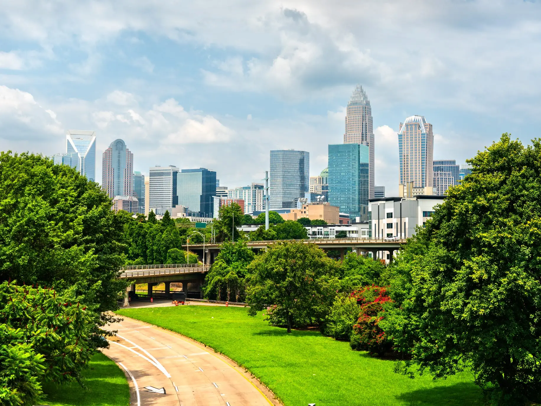 Uptown Charlotte skyline rises beyond green trees, parkland and a curving roadway under a partly cloudy sky.