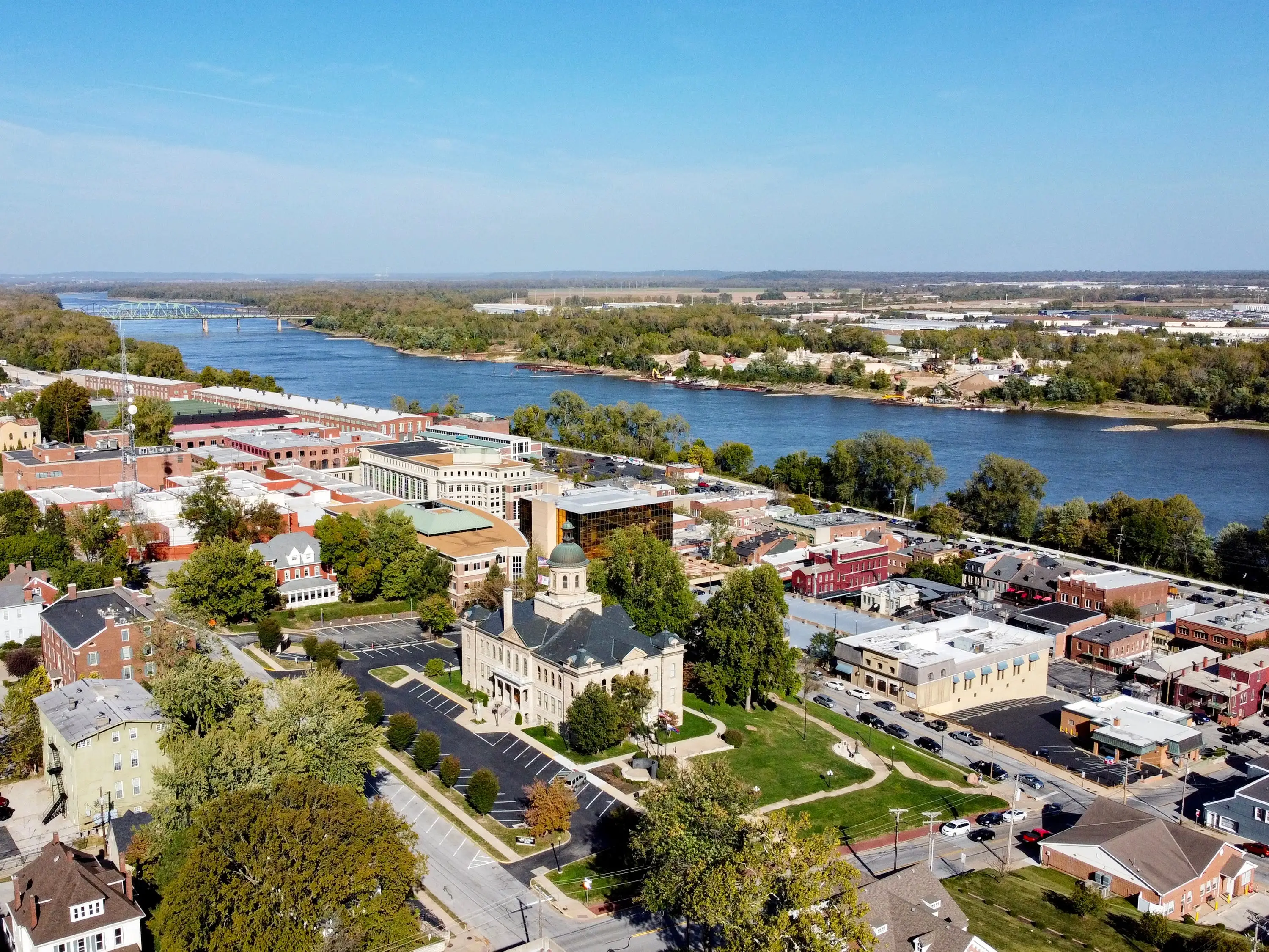Aerial view of a riverside downtown with a domed courthouse, streets, trees, and a broad river under blue sky.