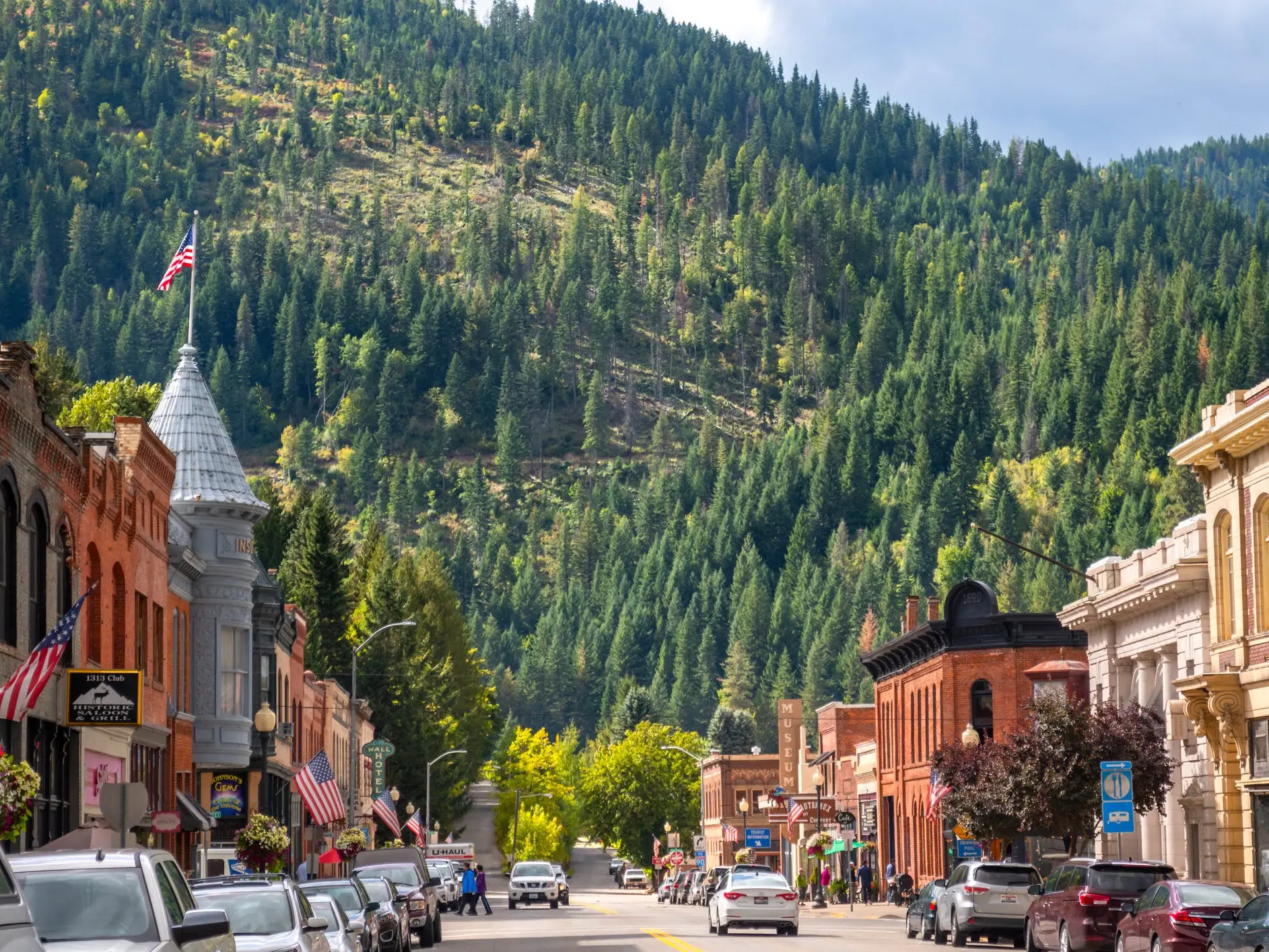 Historic downtown street with American flags, parked cars, and storefronts below a forested mountain.