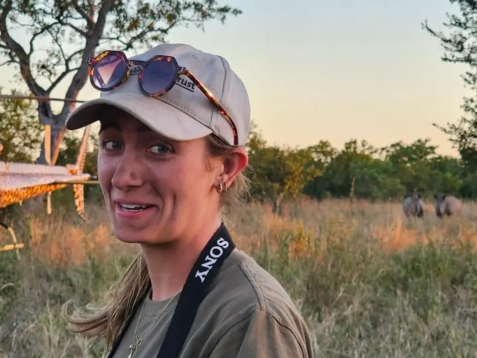 The writer stands in a dry savanna near a helicopter and distant rhinoceroses at sunset in Zimbabwe.