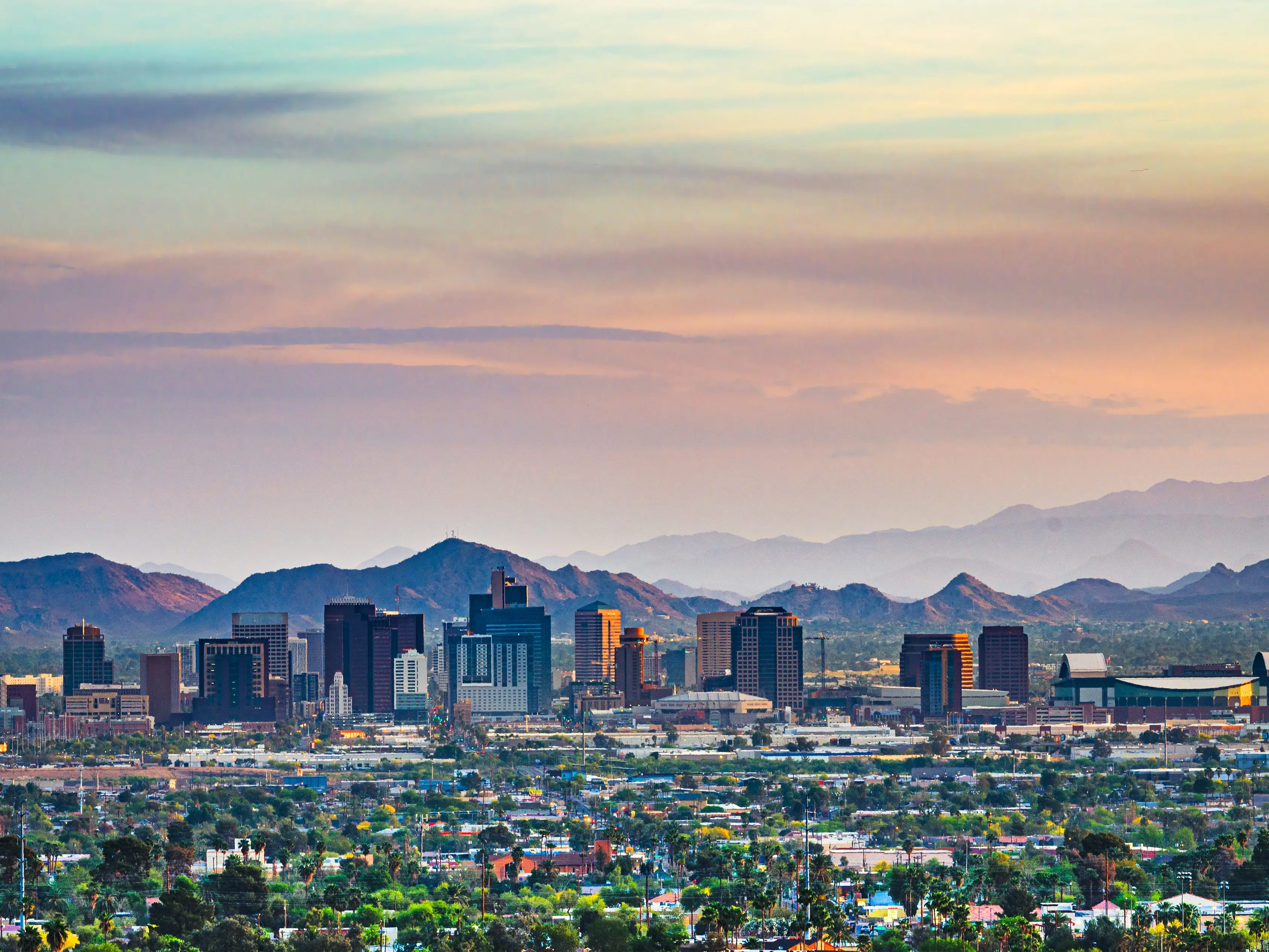 Downtown Phoenix skyline sits below desert mountains under a pastel sunset sky.