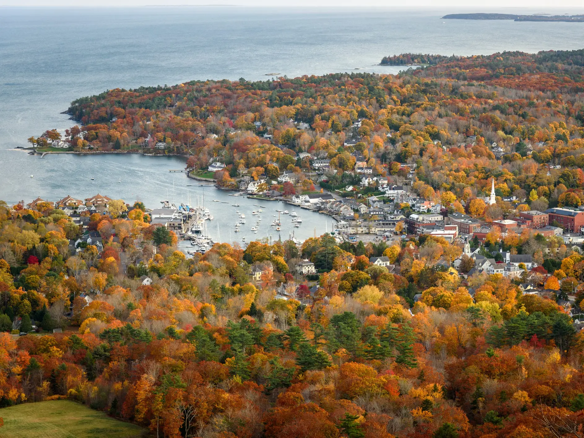 Aerial view of a coastal harbor town surrounded by dense autumn foliage and a marina filled with boats.