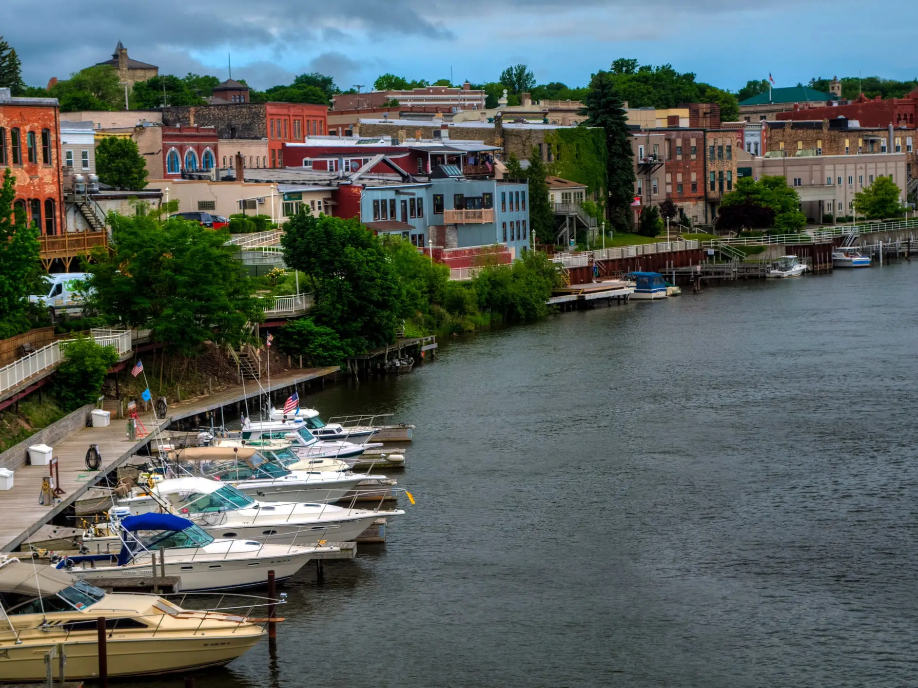 Boats dock along a riverfront marina beside colorful brick buildings under a cloudy sky.